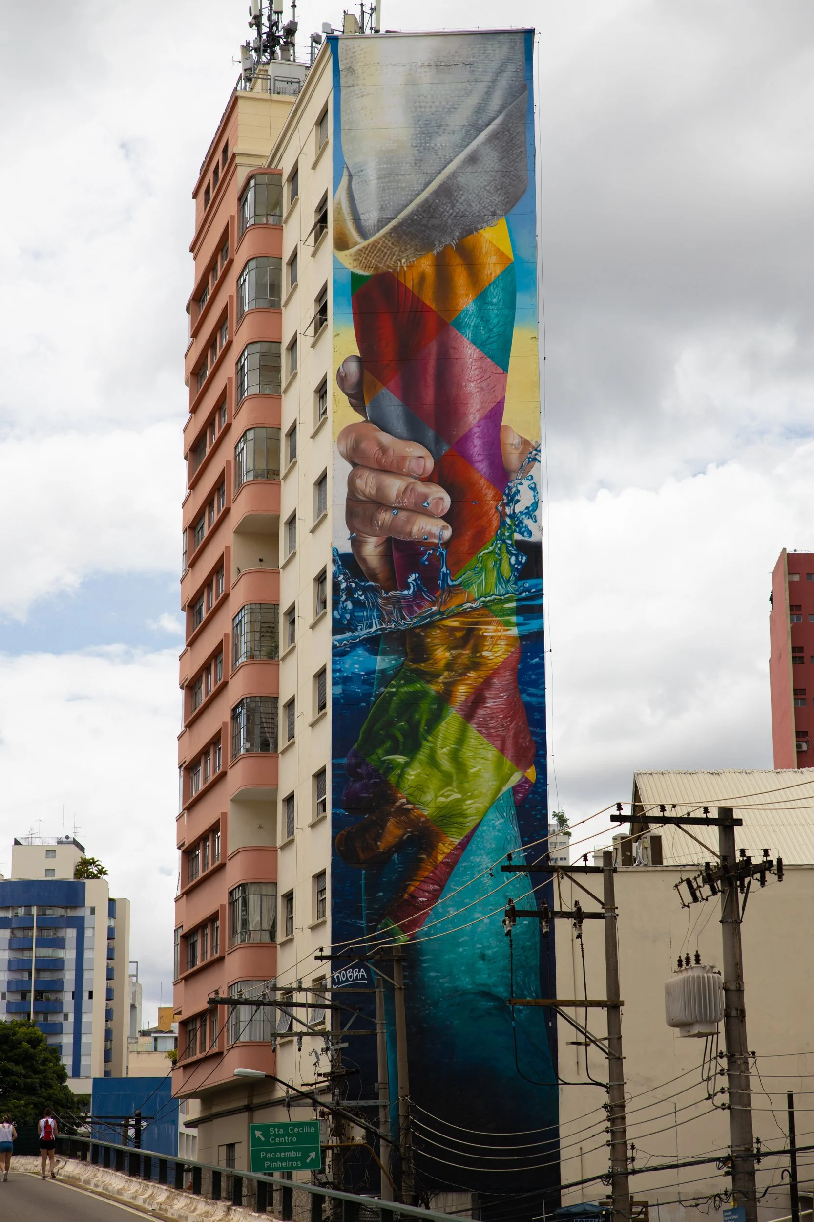 Large colorful mural on the side of a building depicting a hand holding an ice cream cone with a scoop of ice cream, the ice cream appears to blend into a blue water splash at the bottom. The scene is set in an urban area with other high-rise buildin