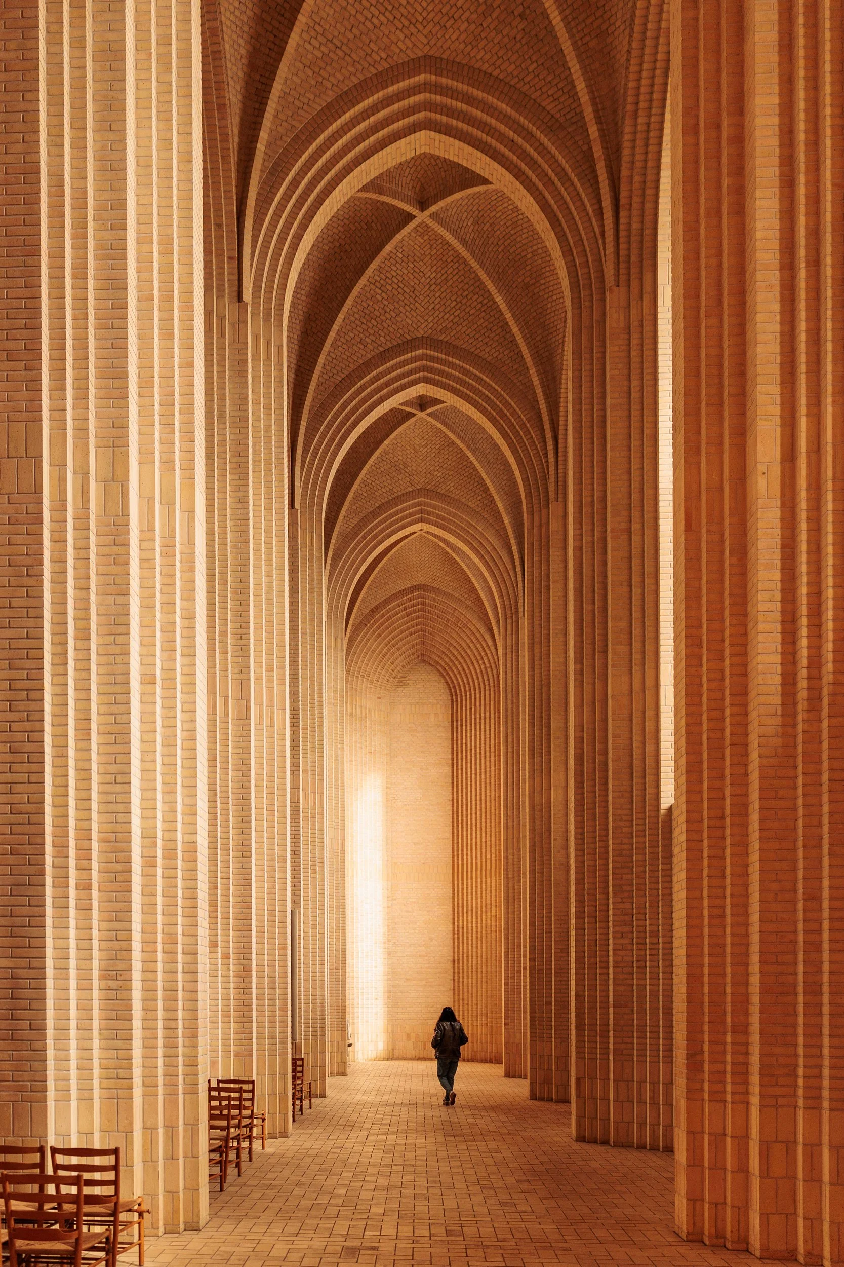 A person walking through a long, arched hallway made of red bricks, with an elegant pattern and lit by soft warm lighting.