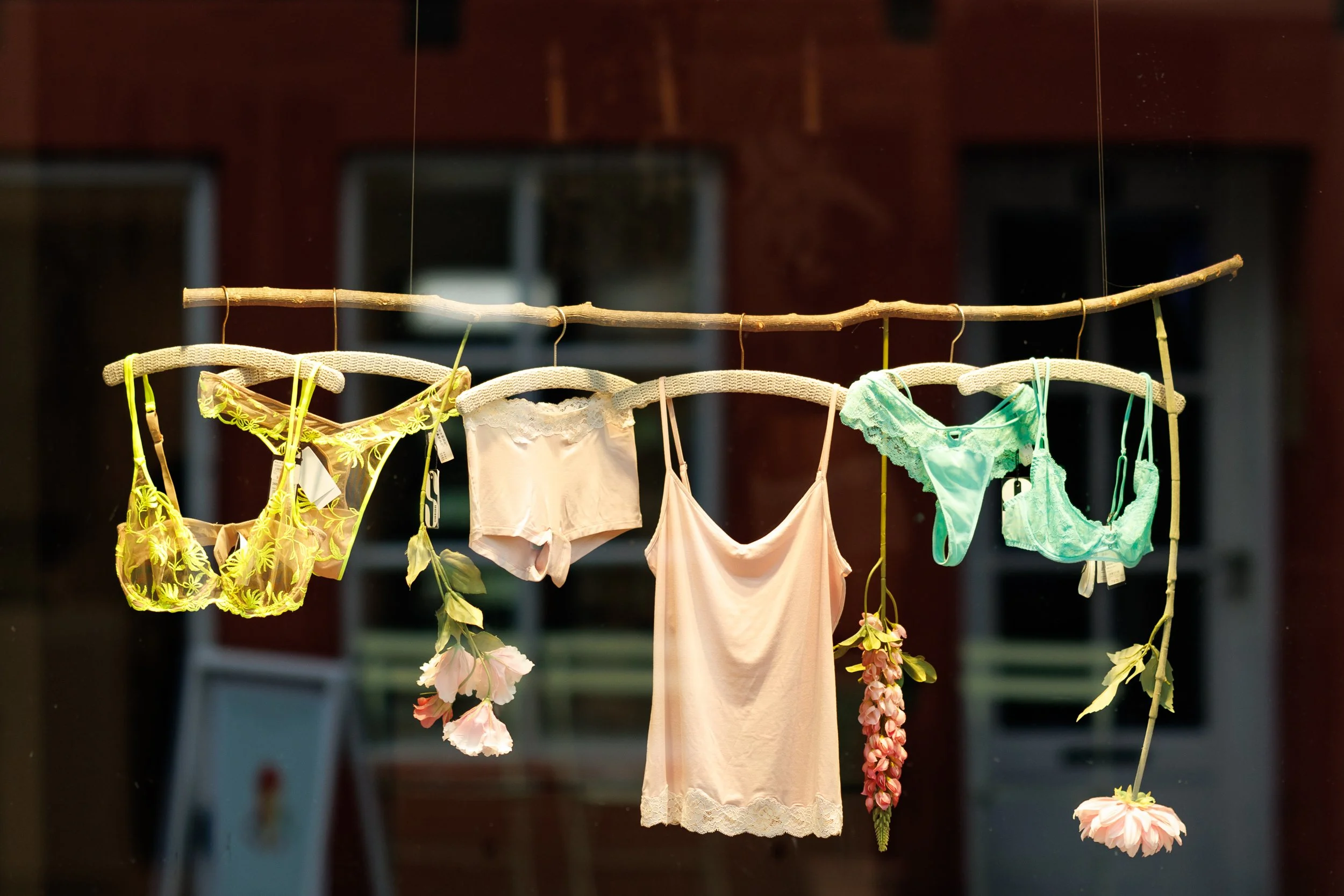 Various women's lingerie and undergarments hanging on a wooden stick with pink flowers hanging beneath, displayed in front of a window.