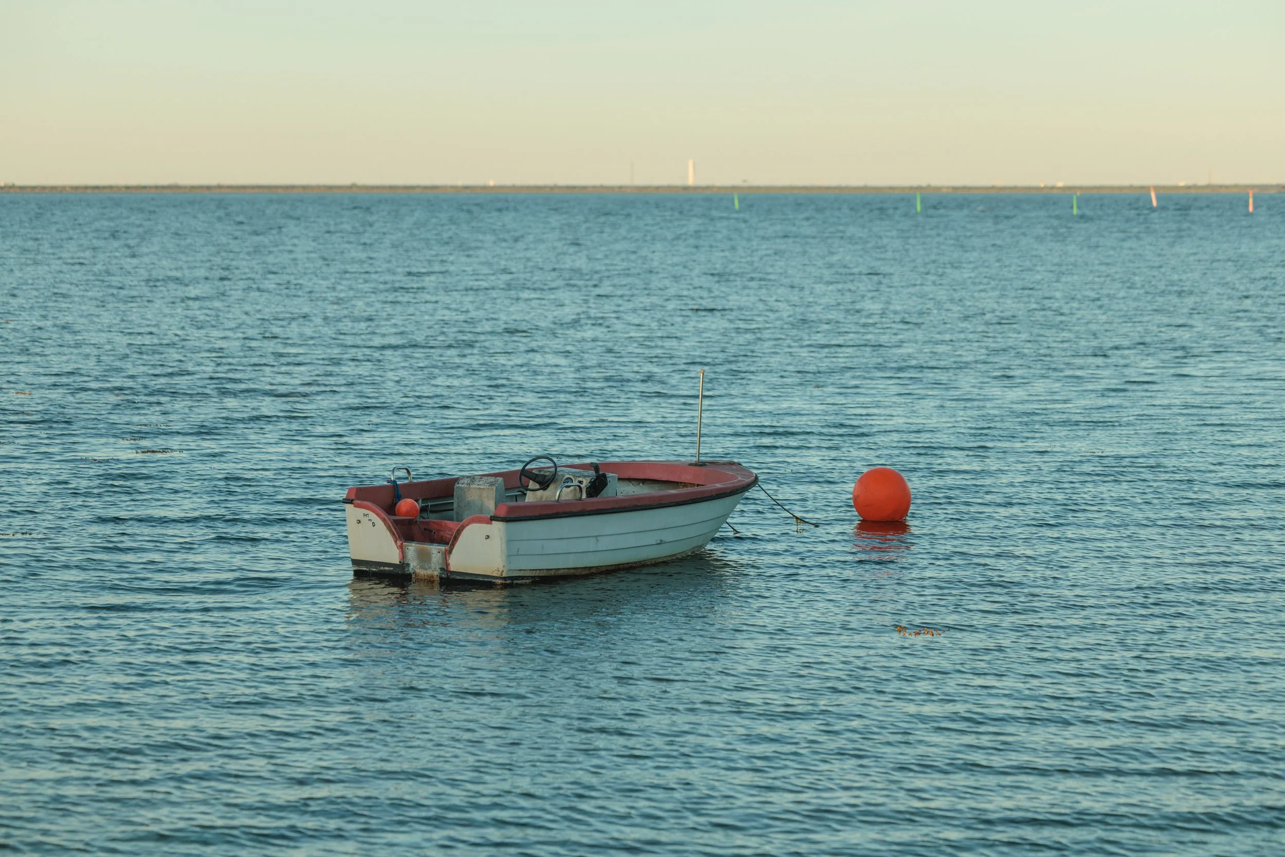 A small boat with a white and red hull floating on calm water near an orange buoy, with a distant horizon and some navigation markers in the water.