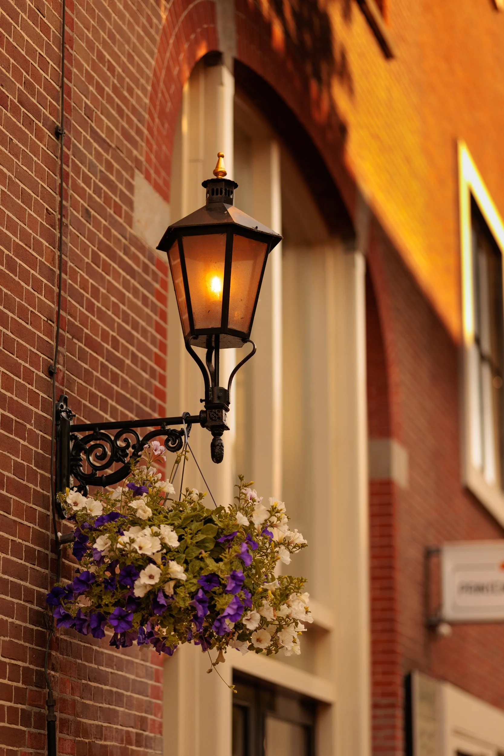 An old-fashioned black lantern with a lit warm yellow light, mounted on a decorative iron bracket beside a brick building. Below the lantern, there is a hanging flower basket with white and purple flowers. The background shows a brick wall with windo