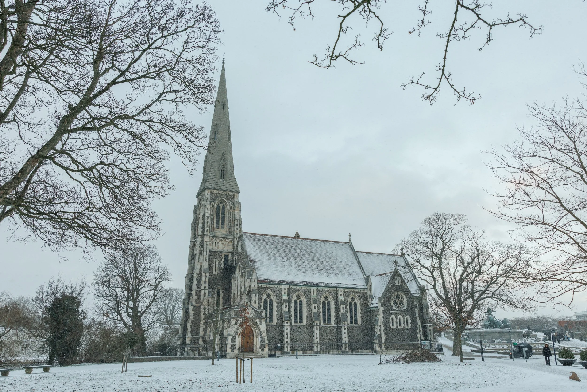A Gothic-style church with a tall steeple, surrounded by leafless trees and snow on the ground on a cloudy winter day.