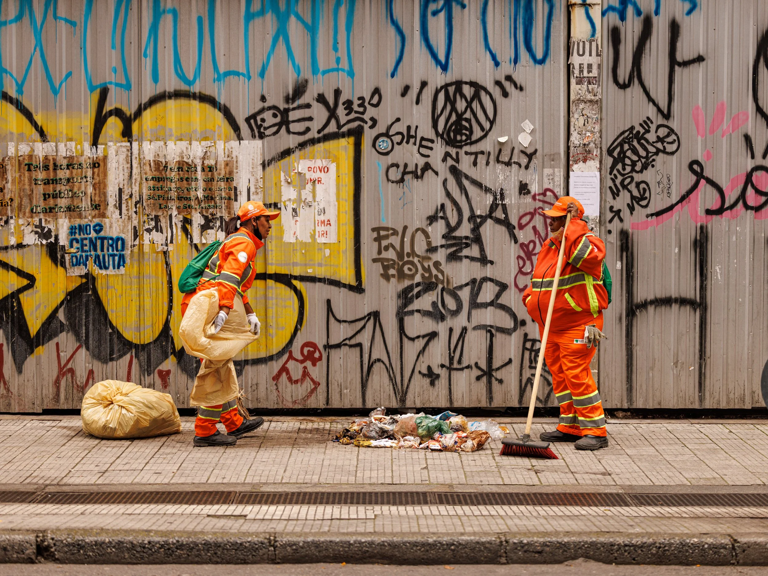 Two sanitation workers in orange uniforms and orange hats stand on the sidewalk near a large pile of trash, with one holding a broom. Behind them is a graffiti-covered metal wall.