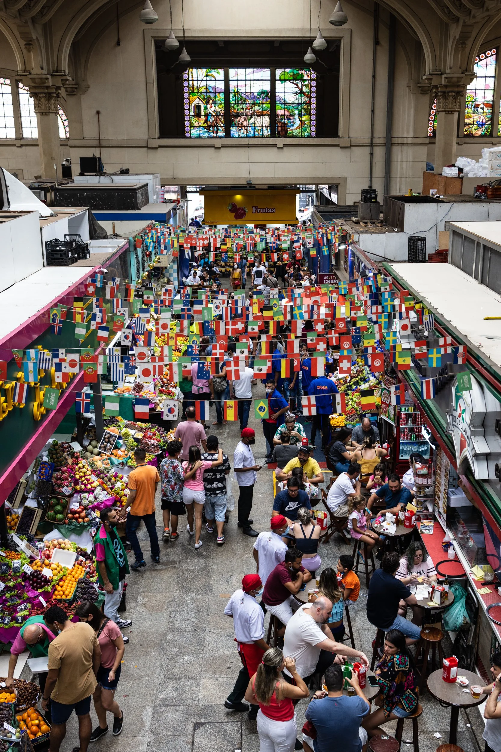 Inside a busy indoor marketplace with stalls selling fruits and vegetables. There are many people shopping, chatting, and sitting at tables. Decorative flags hang above, and stained glass windows depict a pastoral scene.