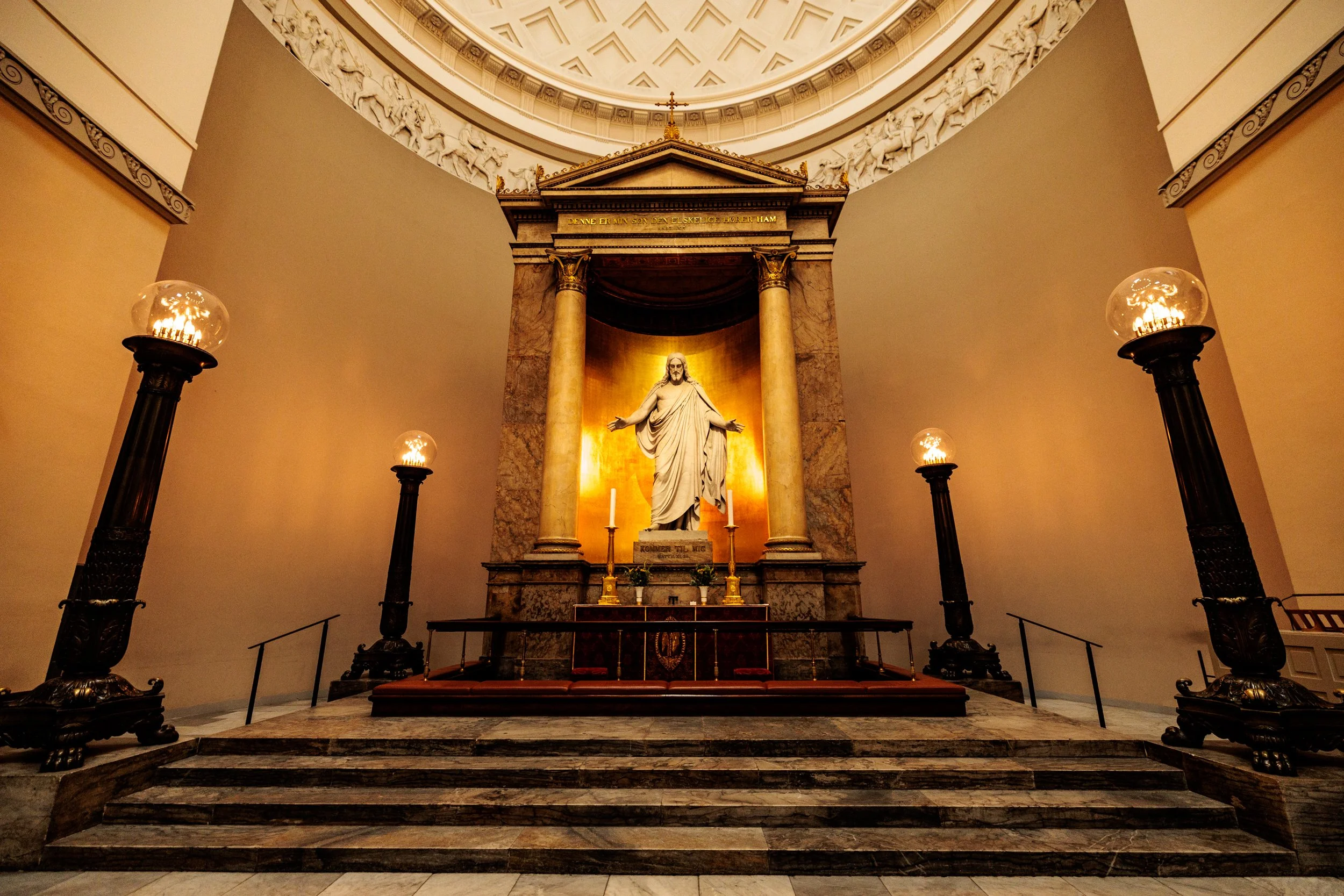 Interior of a church or cathedral altar with a sculpture of Jesus Christ, surrounded by ornate lamps on tall stands and steps leading up to the altar, featuring classical architectural elements and detailed relief sculptures in the ceiling.