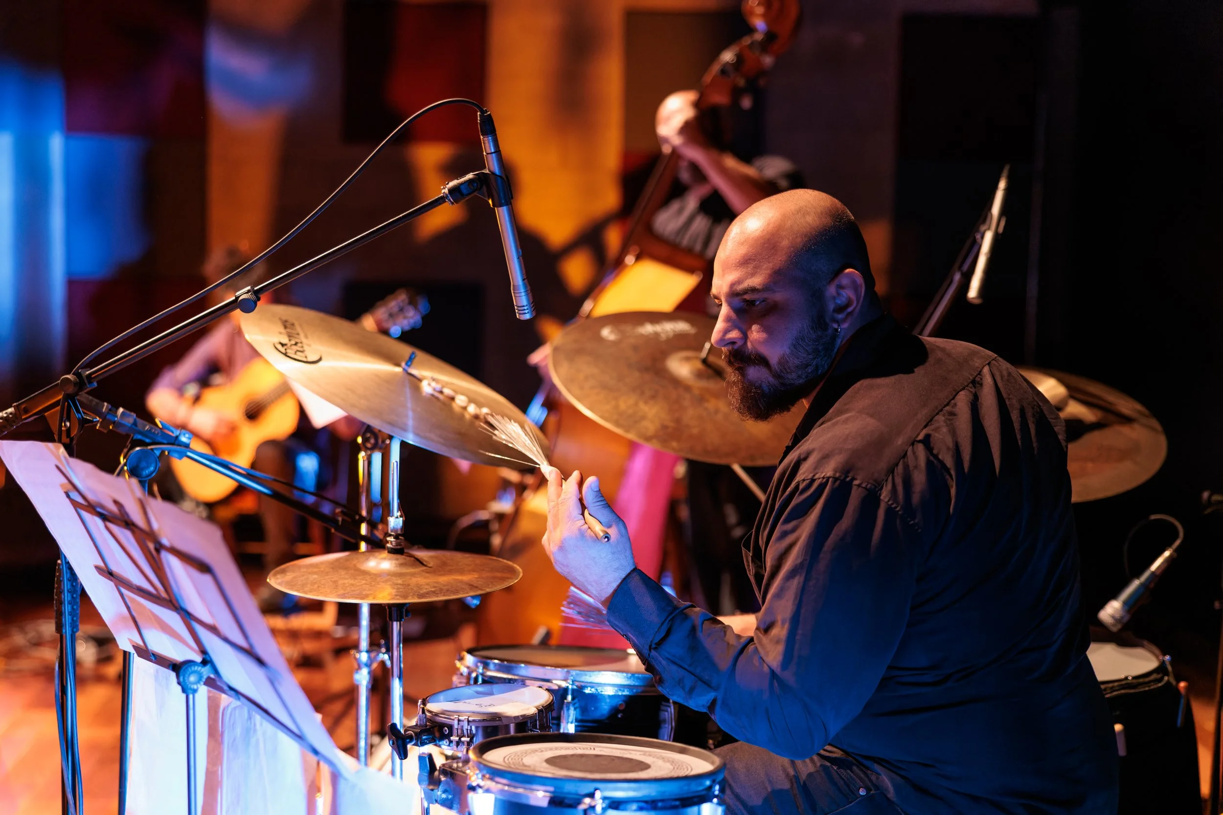 A musician with a shaved head and beard plays drums during a live performance, with a guitar player blurred in the background. The scene is lit with warm lighting in a dark venue.