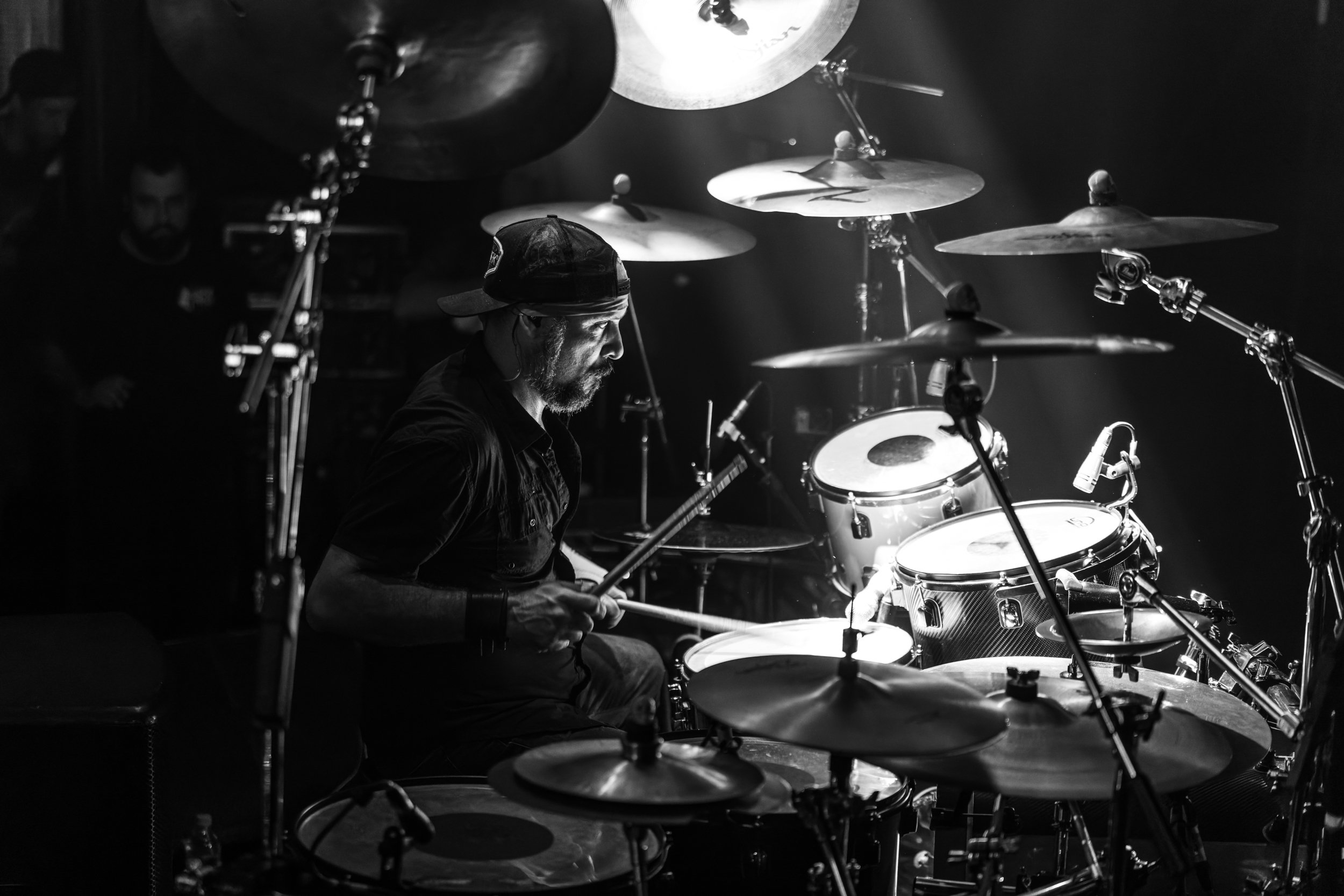 A black and white photo of a drummer wearing a cap, playing his drum set on stage. The scene is lit with spotlights, highlighting his focused expression.