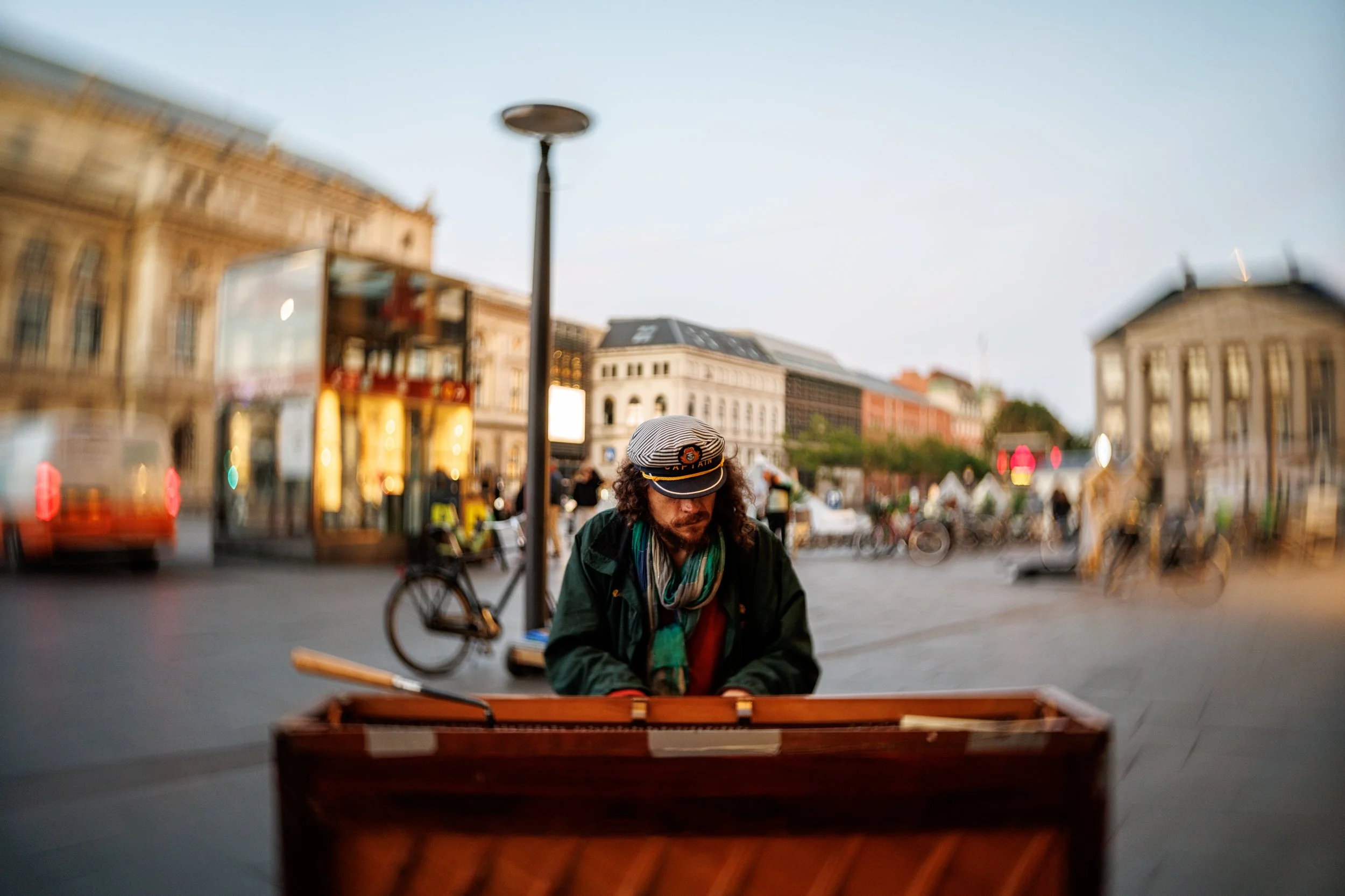 A street singer with long curly hair playing a keyboard outdoors in a city square during dusk, with historic buildings in the background.