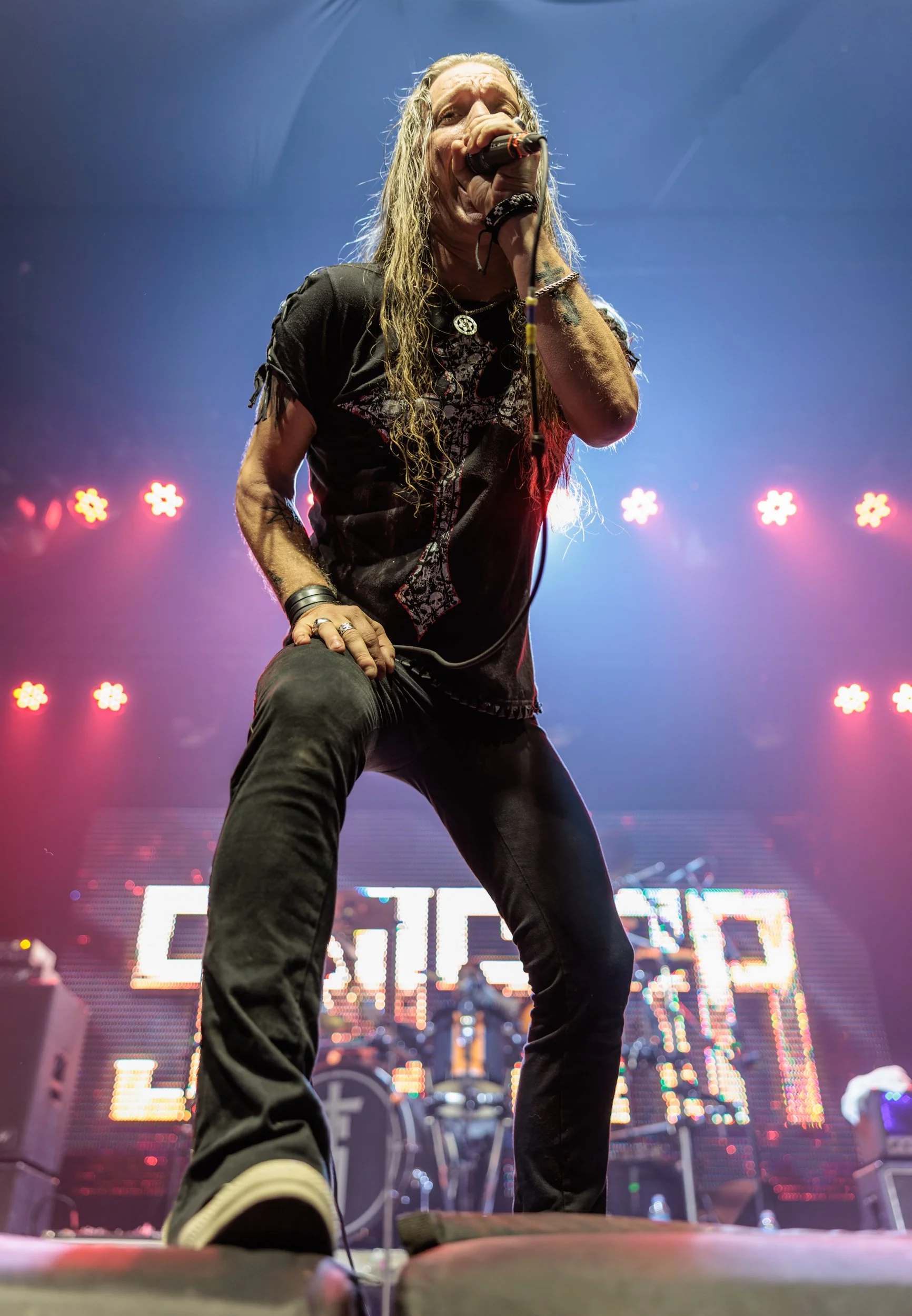 A long-haired male singer holding a microphone on stage during a concert, with colorful stage lights and LED screen in the background.