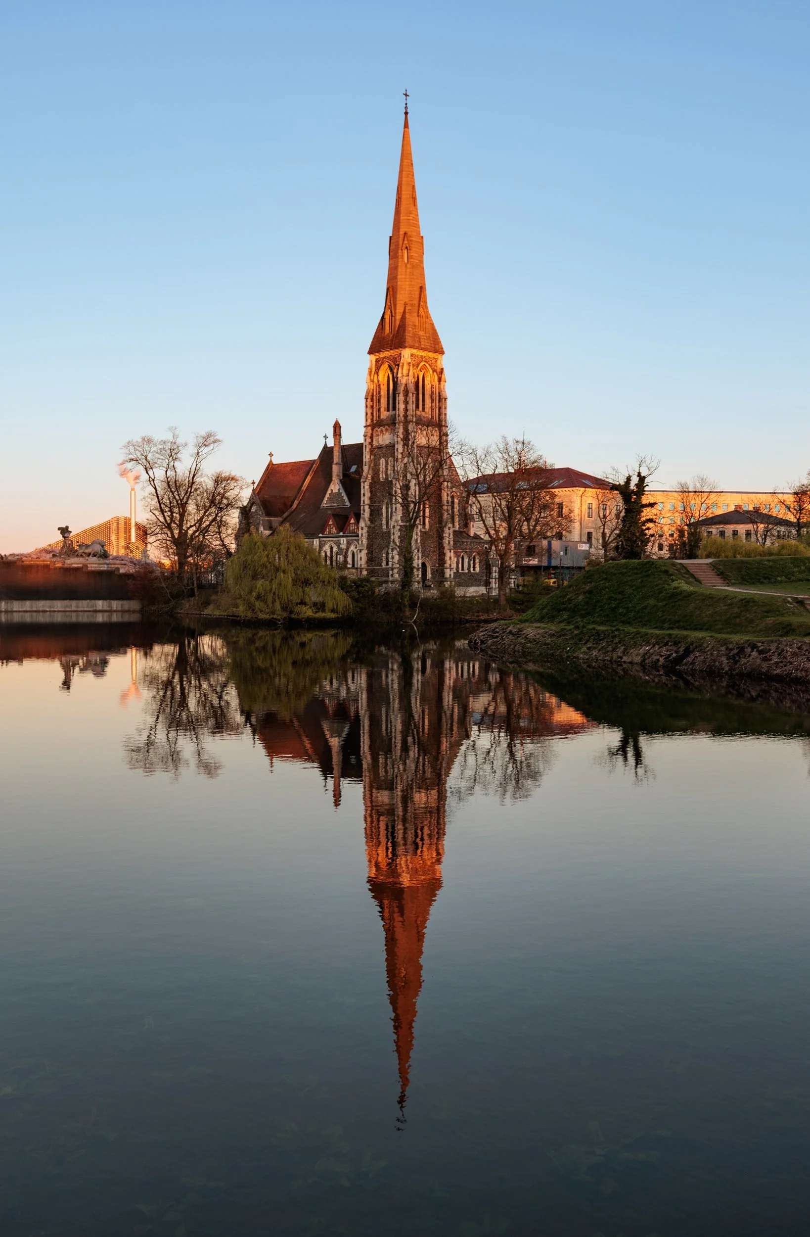 A tall church with a steeple reflected in a calm body of water, surrounded by leafless trees and a few buildings during sunset.