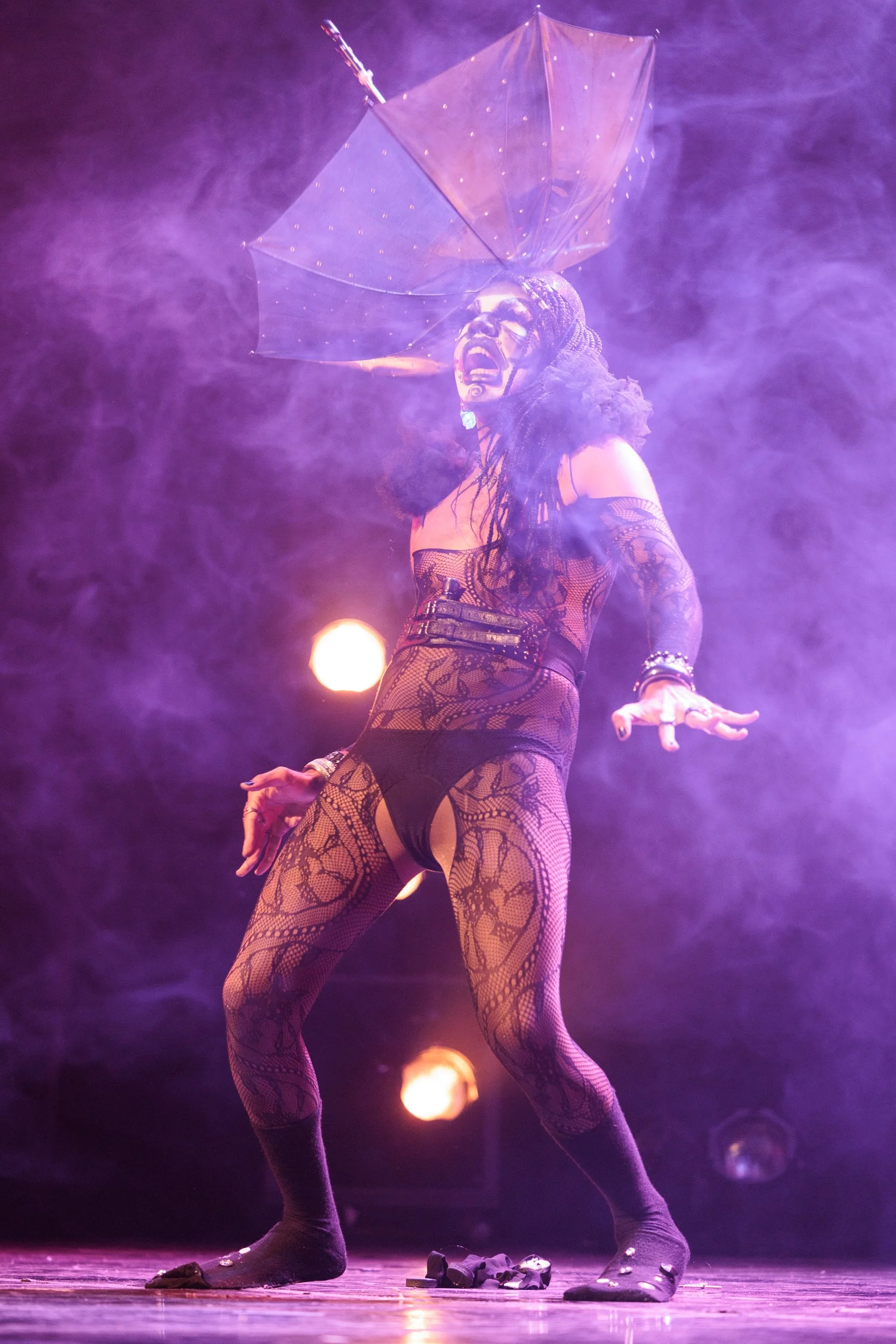 A performer on stage in an elaborate costume, wearing fishnet tights and high-heeled shoes. They hold a black umbrella with light embellishments, with their head arched back and mouth open as if singing or shouting. The stage is filled with purple an