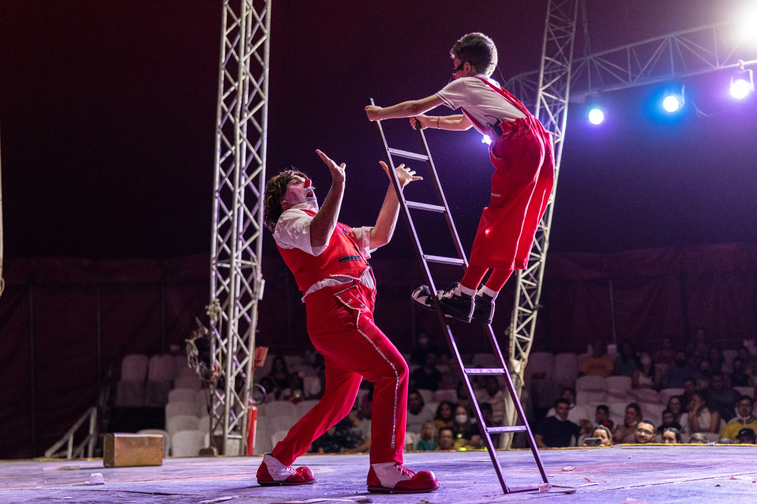 Two performers, dressed as clowns, are on stage with one standing on the ground with hands raised and the other sitting on a ladder. They are surrounded by stage lighting and metal scaffolding, and an audience is visible in the background.