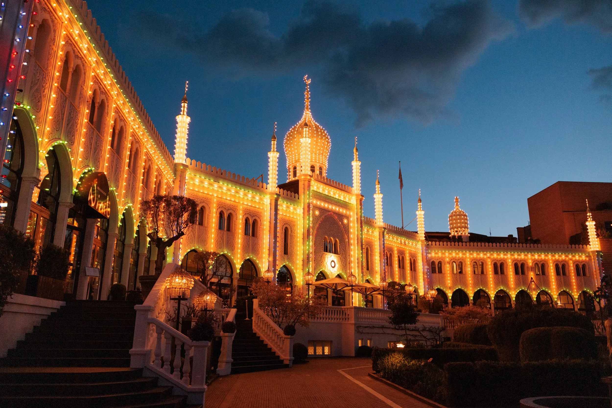 A brightly lit, ornate building with towers and turrets decorated with colorful string lights at night, against a dark blue sky with clouds.