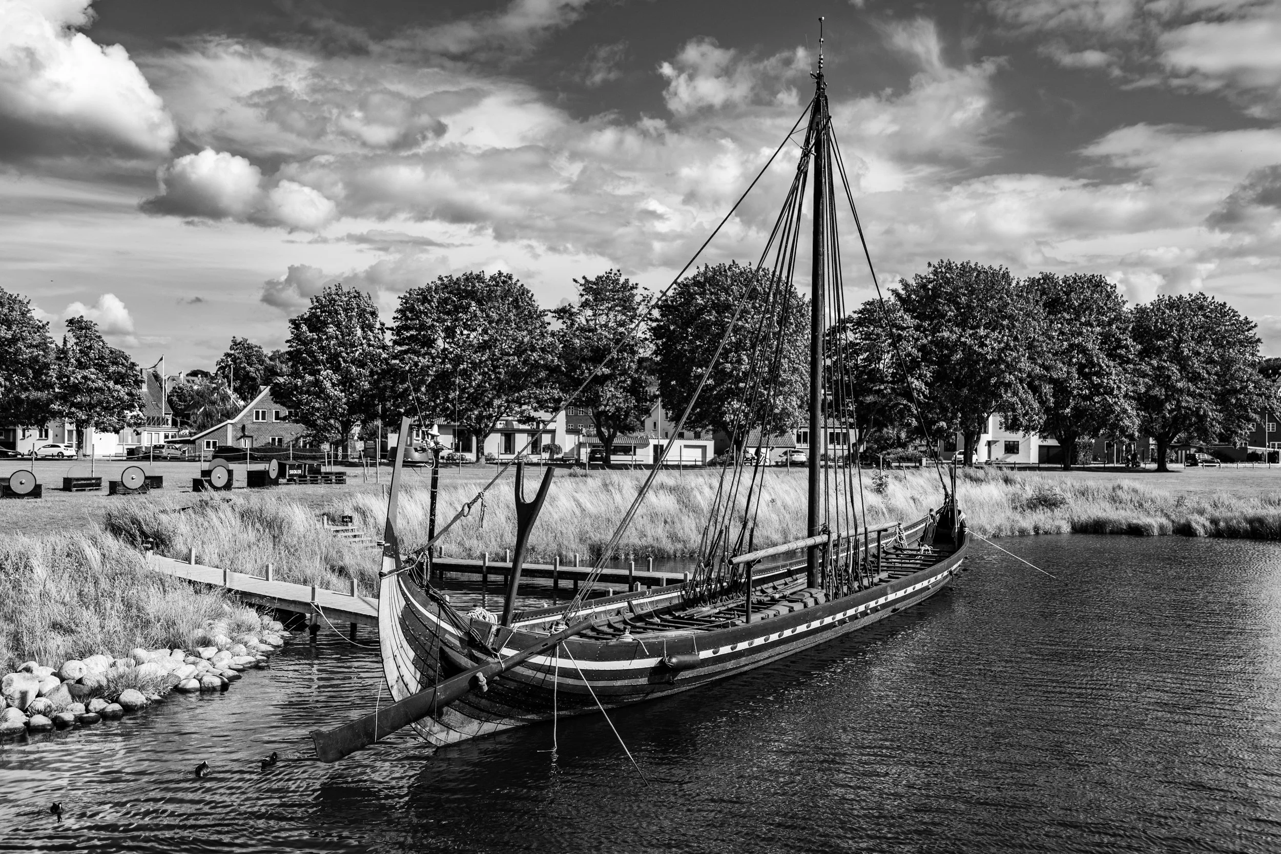 A black and white photograph of a docked boat on a river with houses and trees in the background under a cloudy sky.