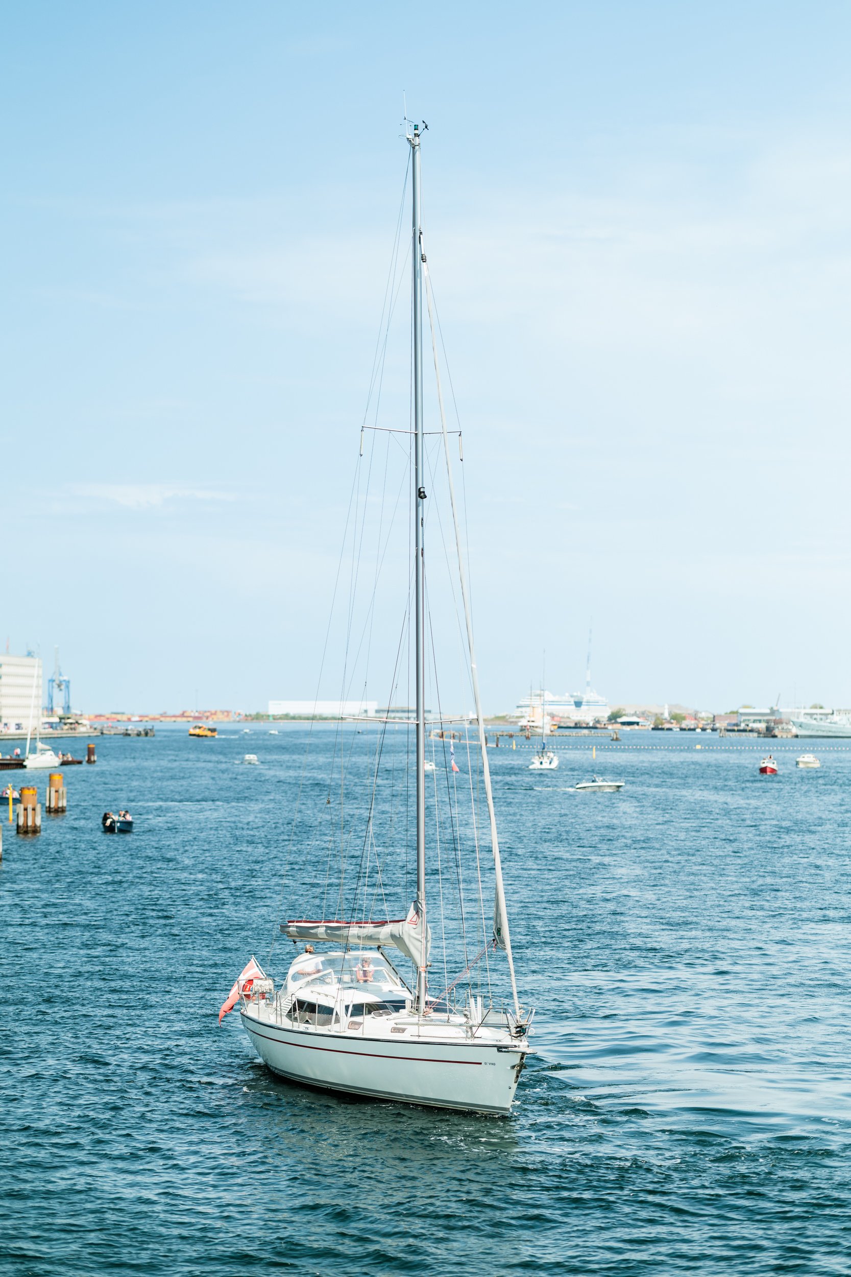 A sailboat with a tall mast sailing in a harbor, with buildings and boats in the background on a sunny day.