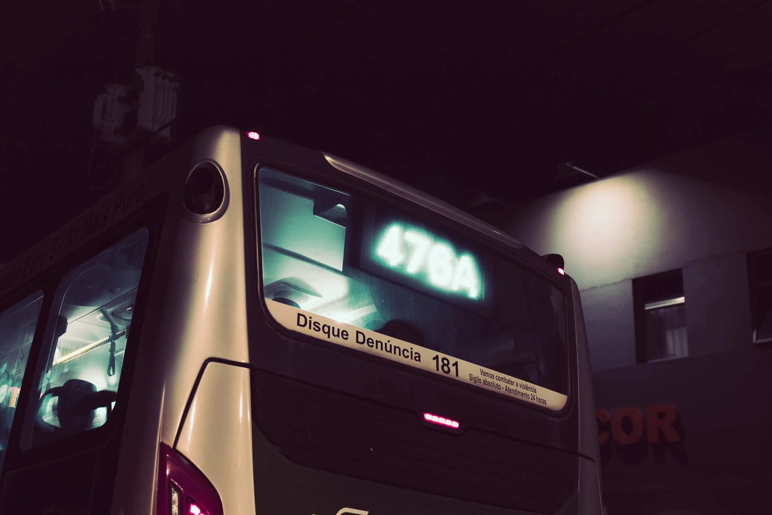 Close-up of a bus's front with a sign that reads 'Disque Denúncia 181' displayed on the windshield, dark environment, and some lights visible.