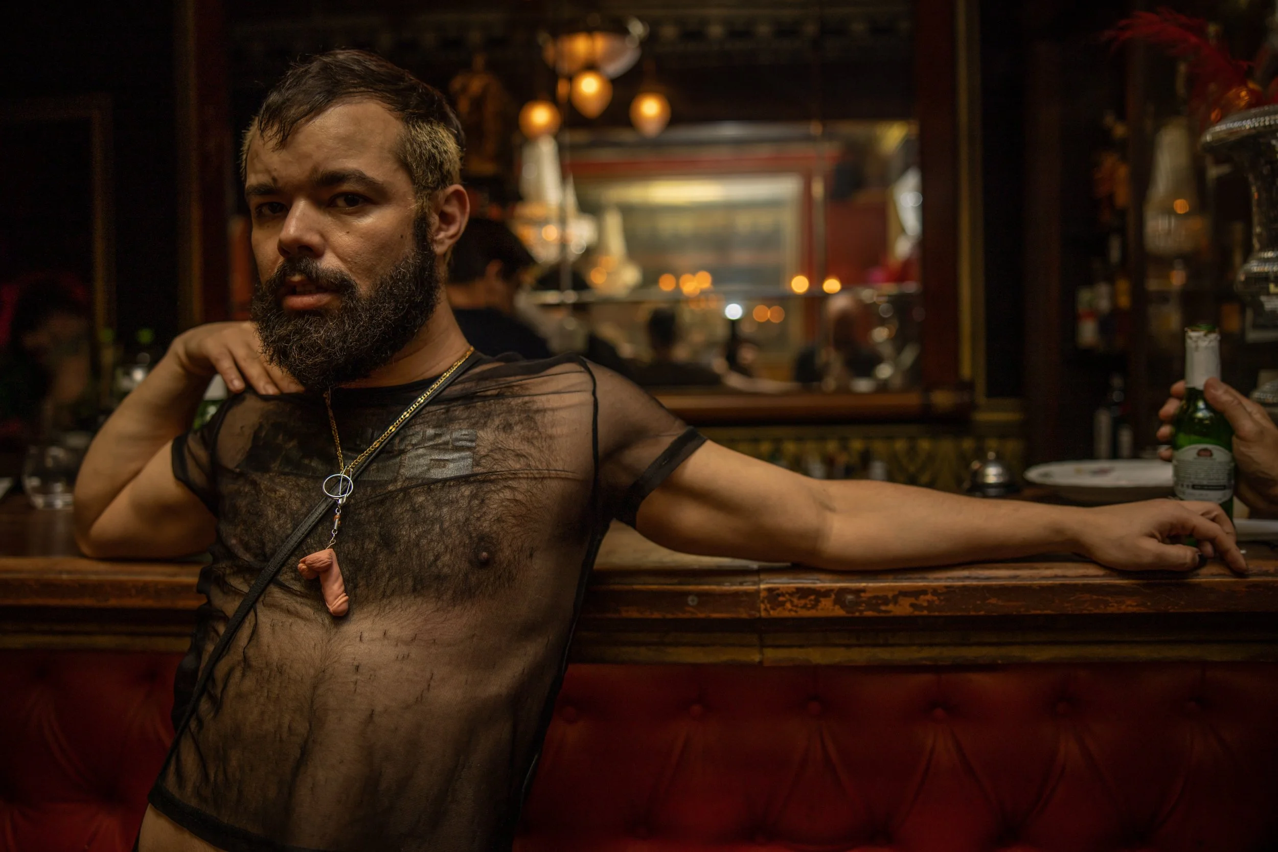 A man with a beard wearing a sheer black mesh shirt and a gold necklace with a phallic pendant, sitting at a bar with a hand on his shoulder, holding a beer bottle in his other hand. The bar has a dark, warm ambiance with hanging lights and blurred b
