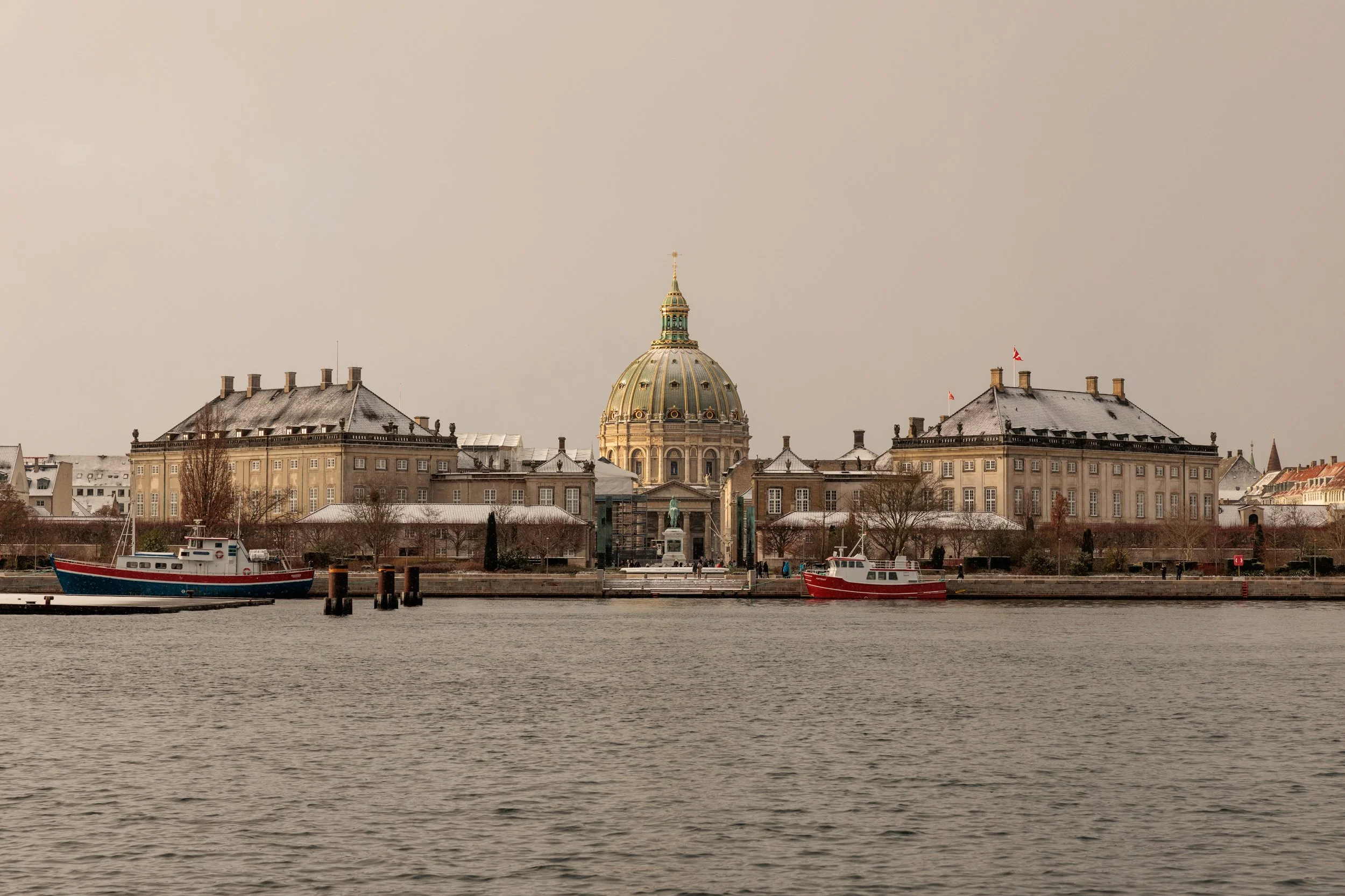 View of a historic building with a large dome and smaller structures, a waterfront with boats, and a cloudy sky.