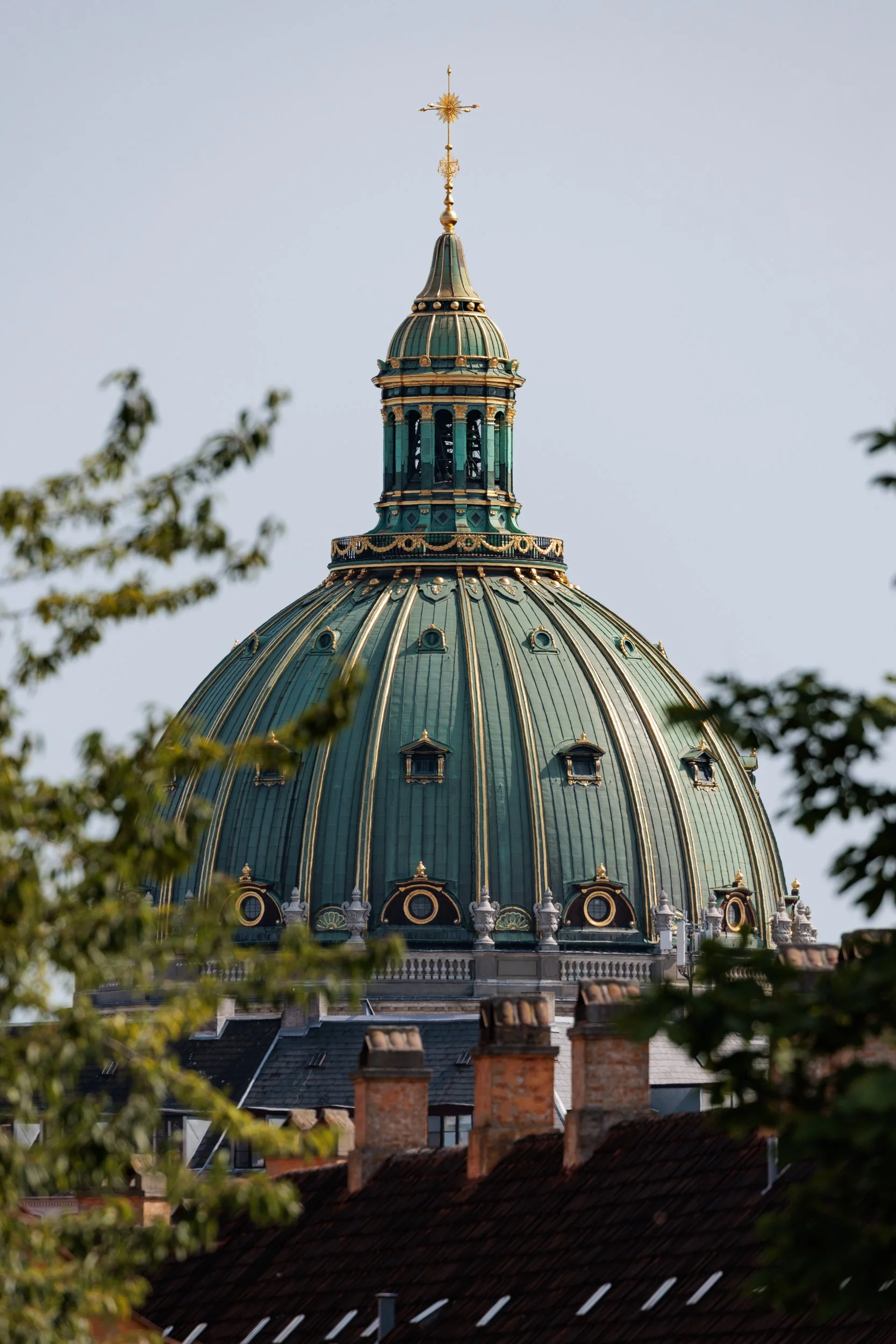 A green ornate dome with gold accents and a gold cross on top, seen through trees, atop a building with chimneys, under a clear sky.