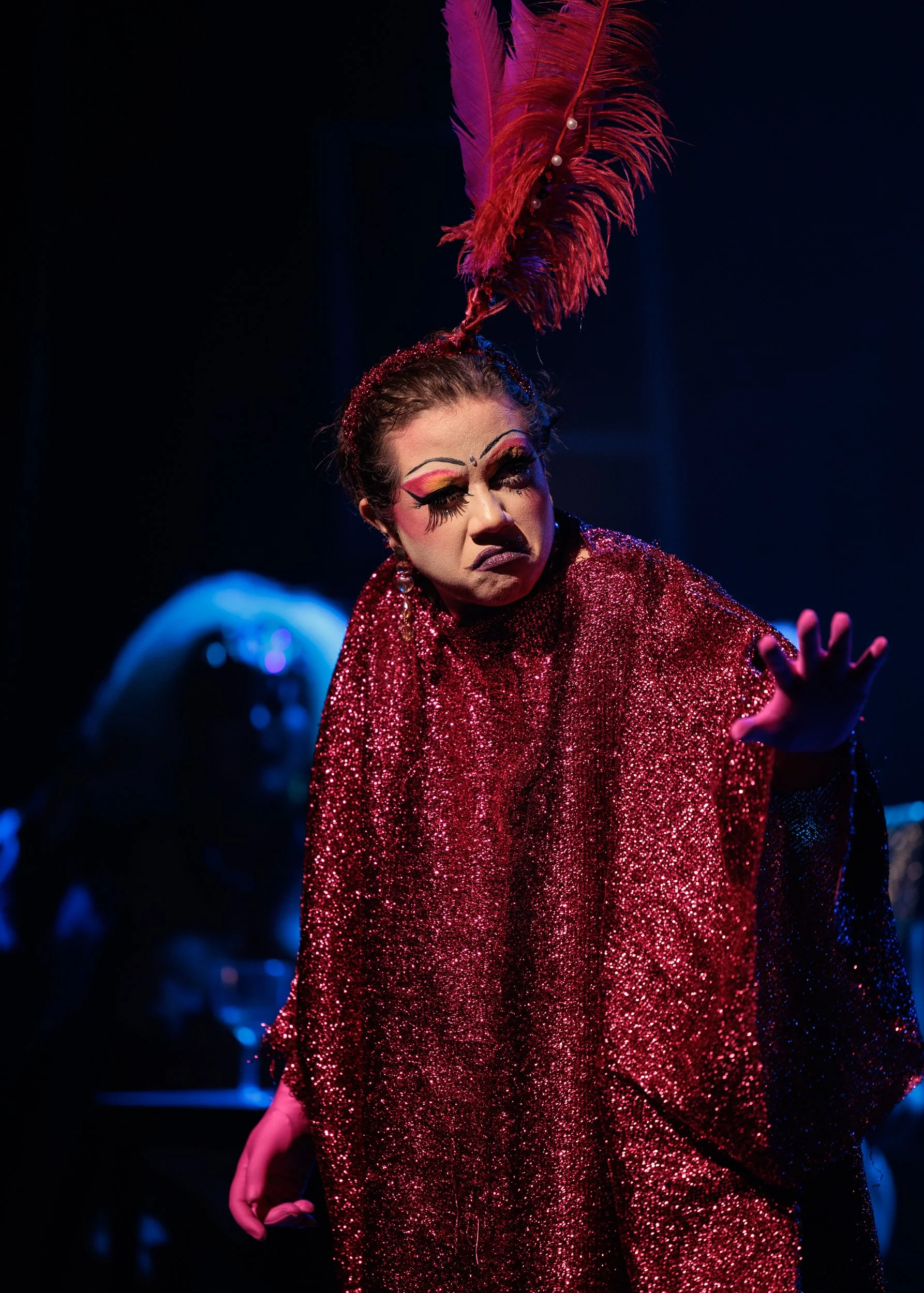 Performer dressed in a red, sparkling costume with large red feathers and pearls, posing with one finger extended and a serious expression on a dark stage.