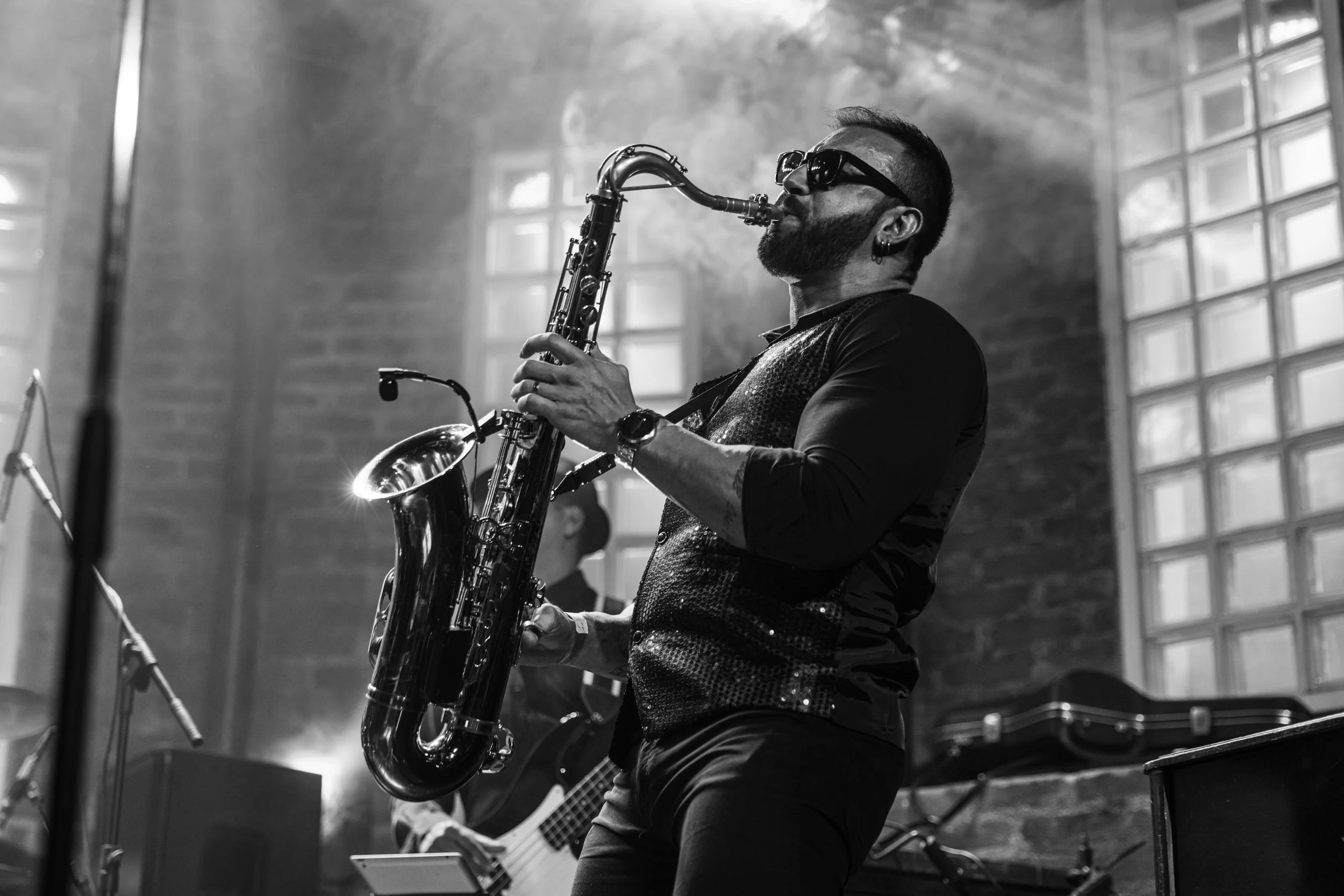 A musician playing a saxophone during a live performance in a dimly lit indoor venue, with a brick wall and windows in the background.