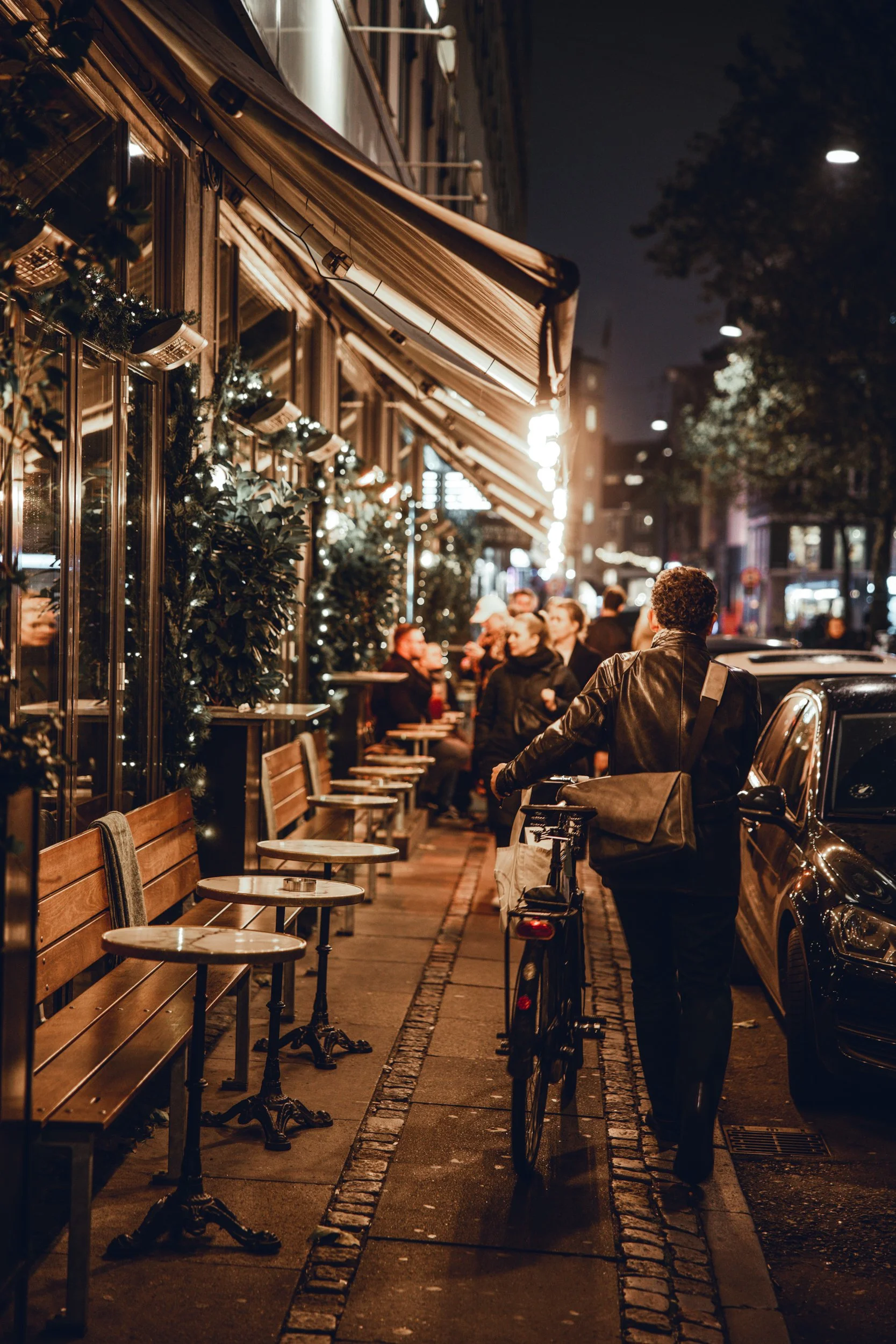 Nighttime street scene with outdoor dining area decorated with Christmas lights, showing a man walking his bicycle along the sidewalk, and people sitting at tables under warm lighting.