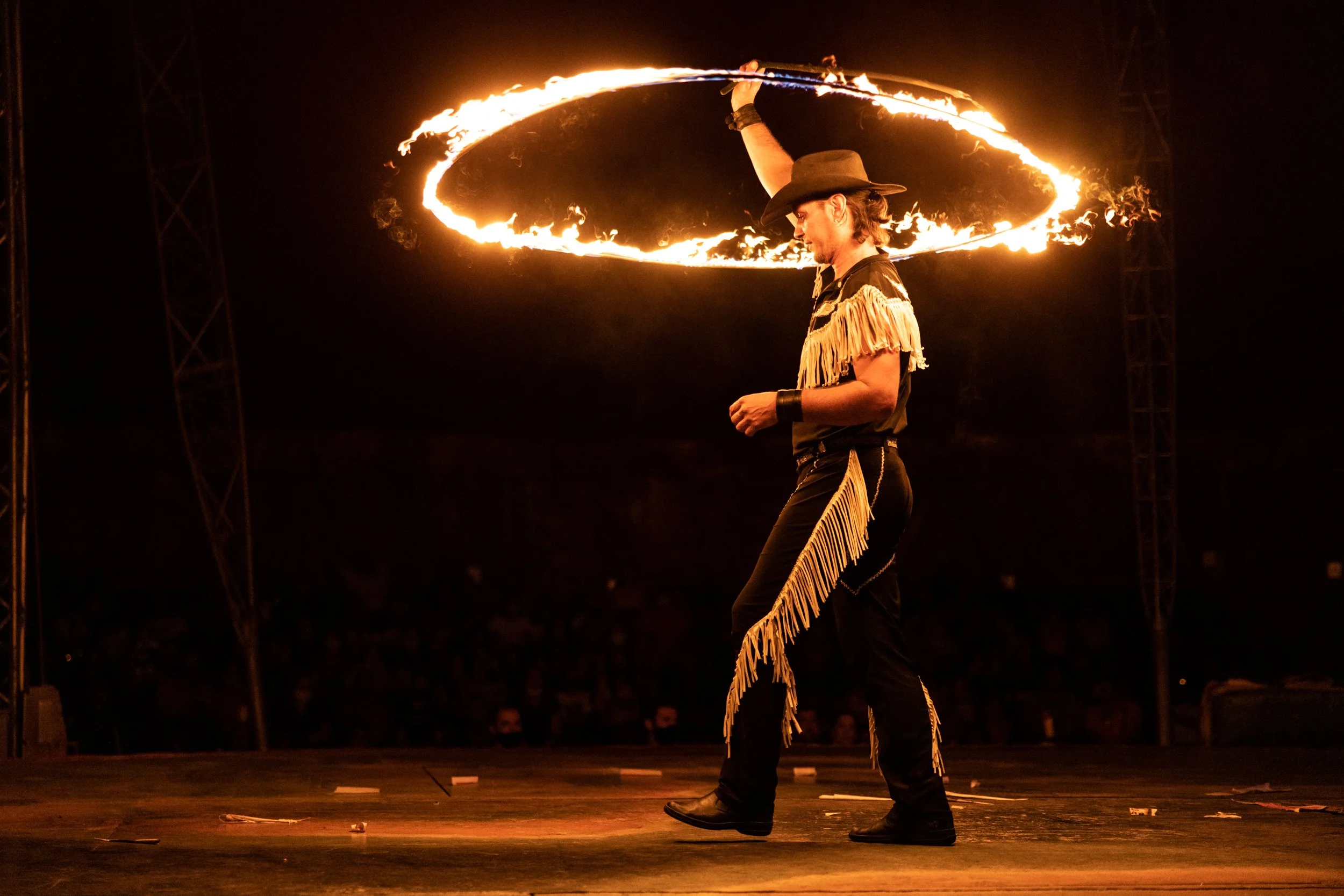 A man dressed in western attire, including a cowboy hat and fringed clothing, performing a fire spinning trick with a flaming hula hoop at night.