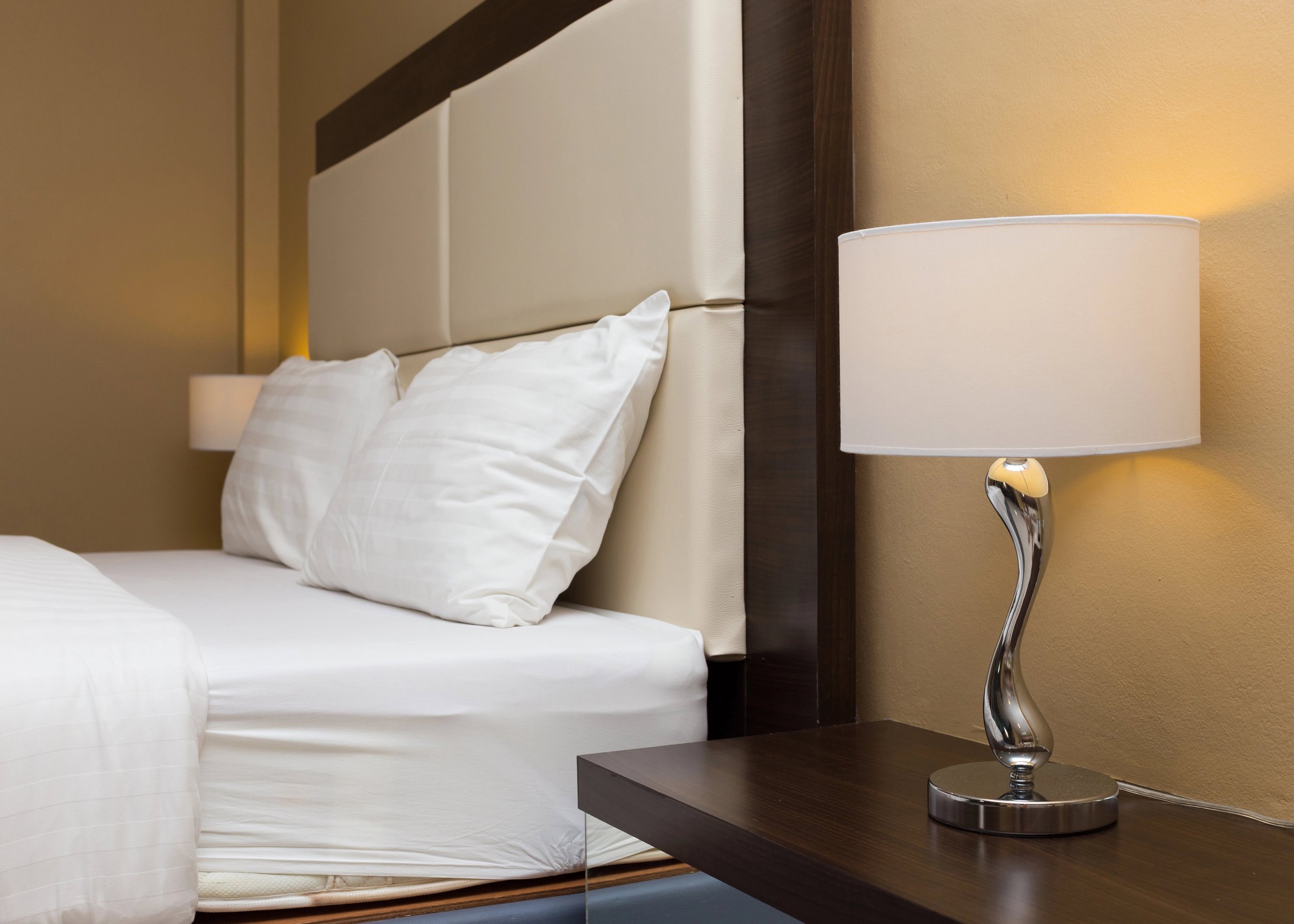 Hotel room close-up of a modern table lamp with a white shade, placed on a dark wooden bedside table next to a bed with white linens and pillows. The background wall is painted a warm beige color.
