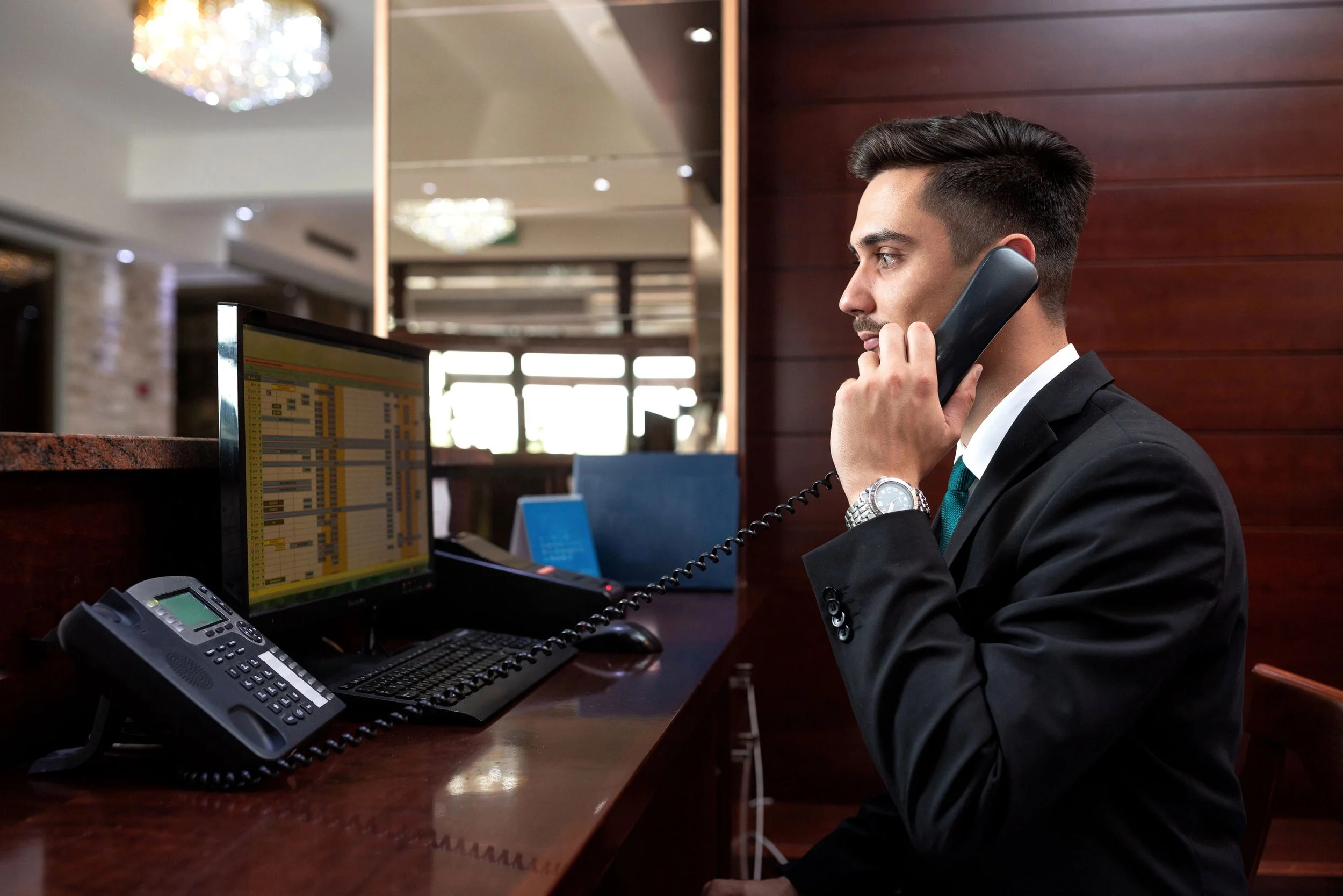 A male hotel concierge in a business suit sits at a desk, using a telephone, in an office lobby with chandeliers and wooden wall panels.