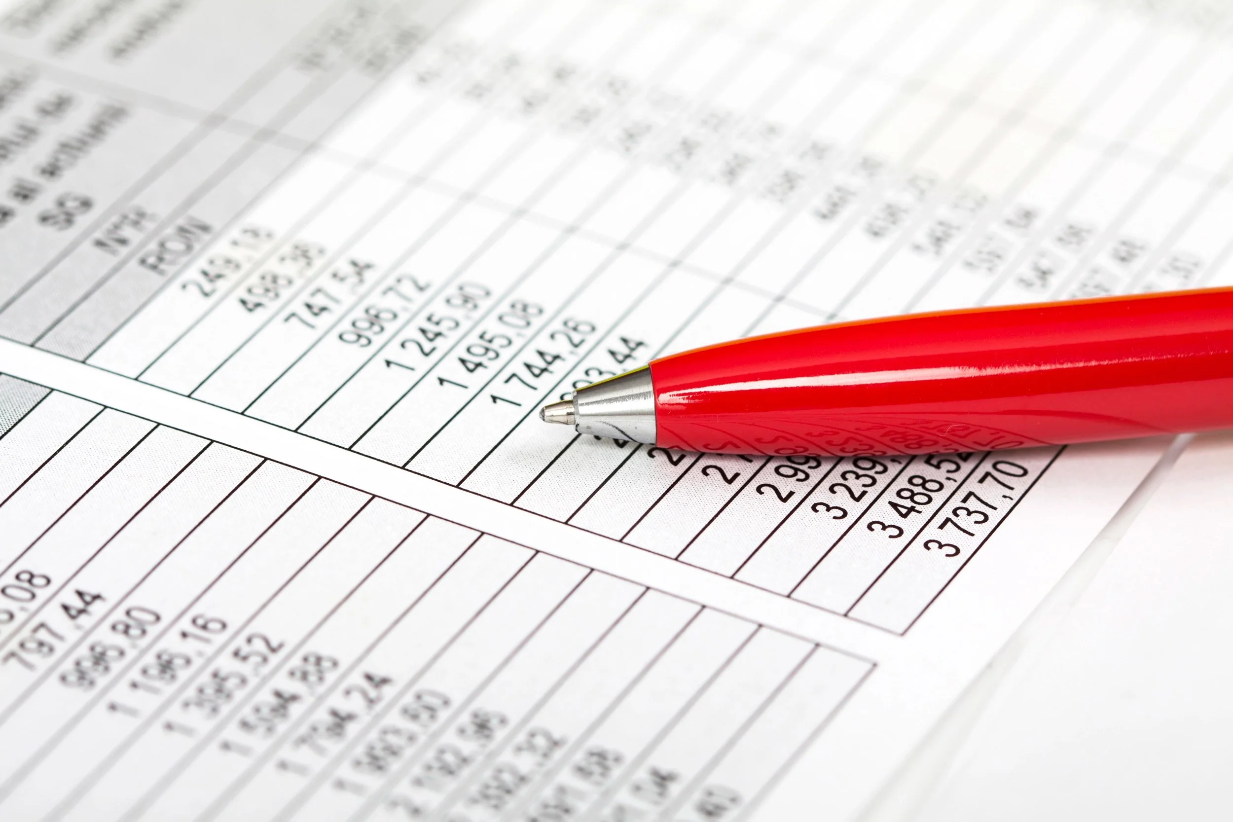 A red pen resting on a printed spreadsheet or ledger with rows and columns of numerical data.