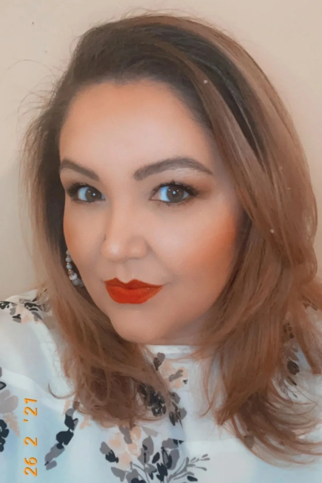 Close-up of a woman with shoulder-length wavy hair, blue eyes, red lipstick, wearing pearl earrings, and a white blouse with black and beige floral pattern.