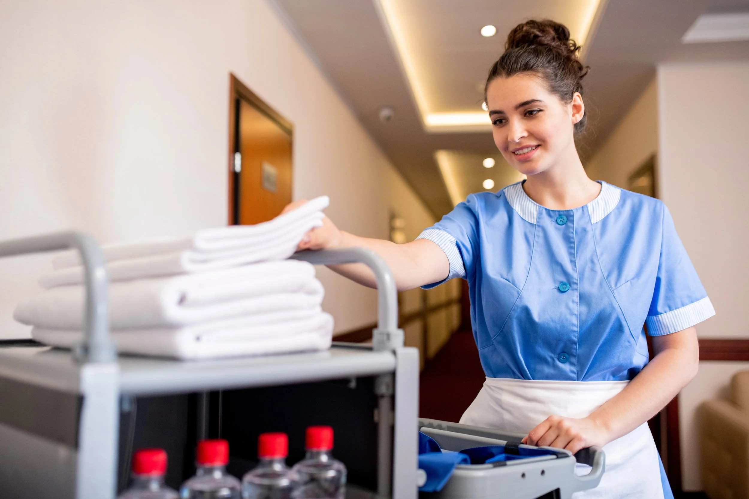 A young female in a blue hotel staff uniform with white accents is placing folded towels on a cart in a clinical setting with warm lighting and a hallway background.