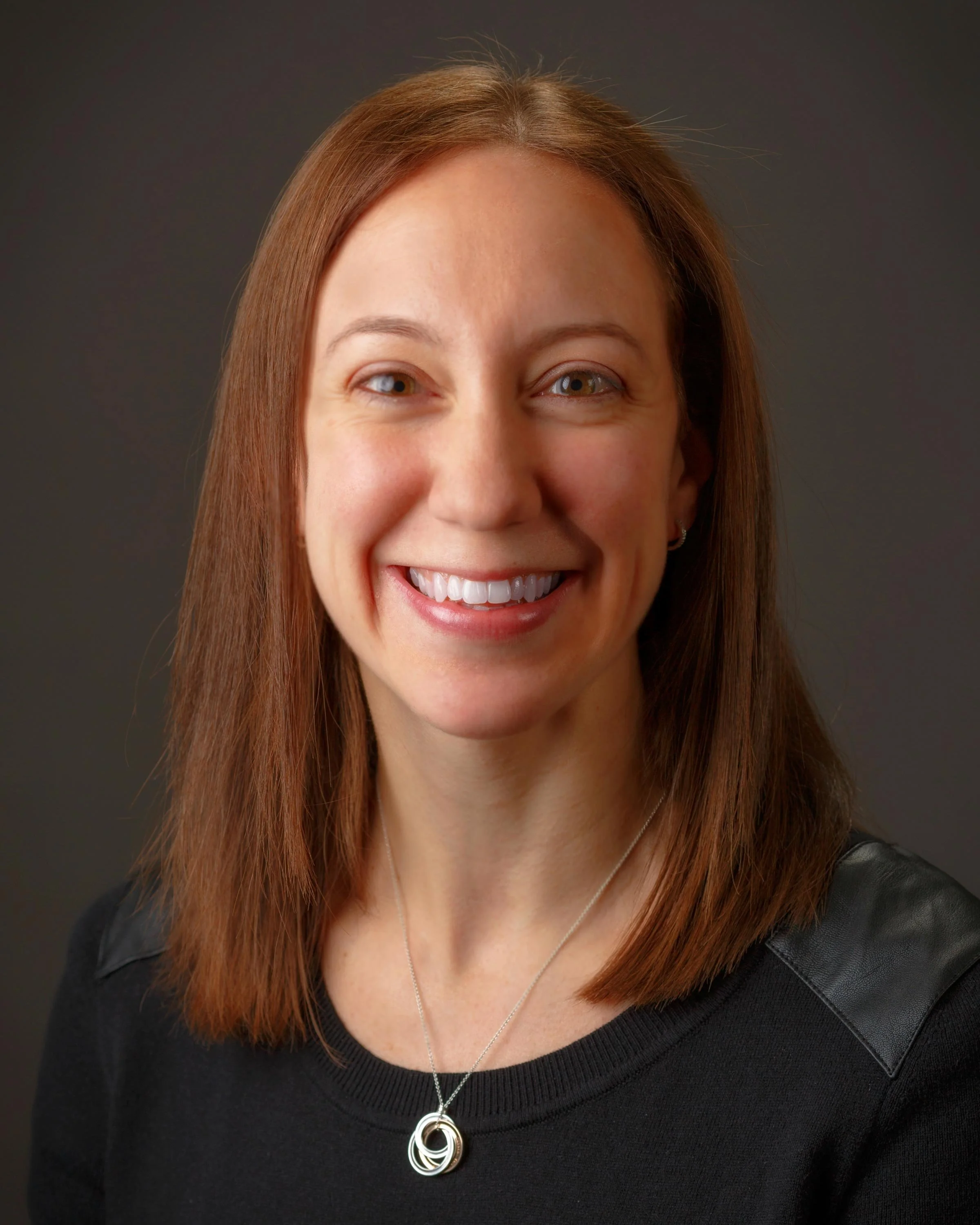 Portrait of Rosemont Project Management's Maricela Vargas, Associate Vice President of Procurement. A woman with shoulder-length brown hair wearing a black top and a necklace against a dark background.