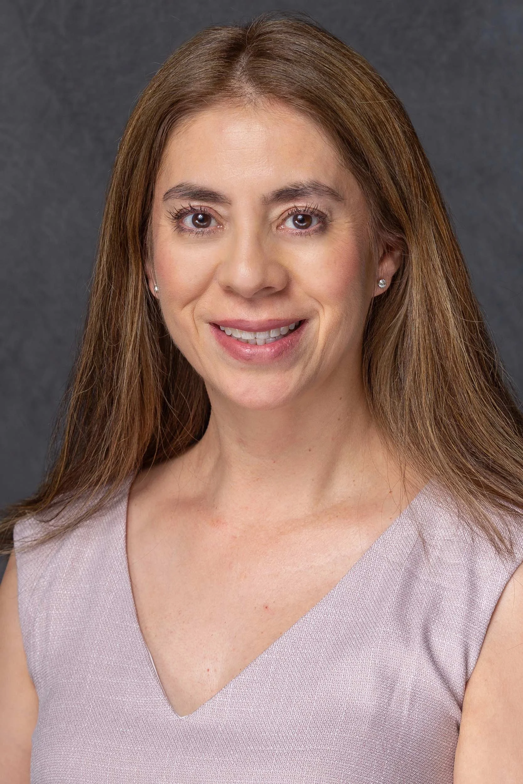 A headshot of a Maricela Vegas with long brown hair, smiling, wearing a sleeveless light pink top, against a dark gray background.