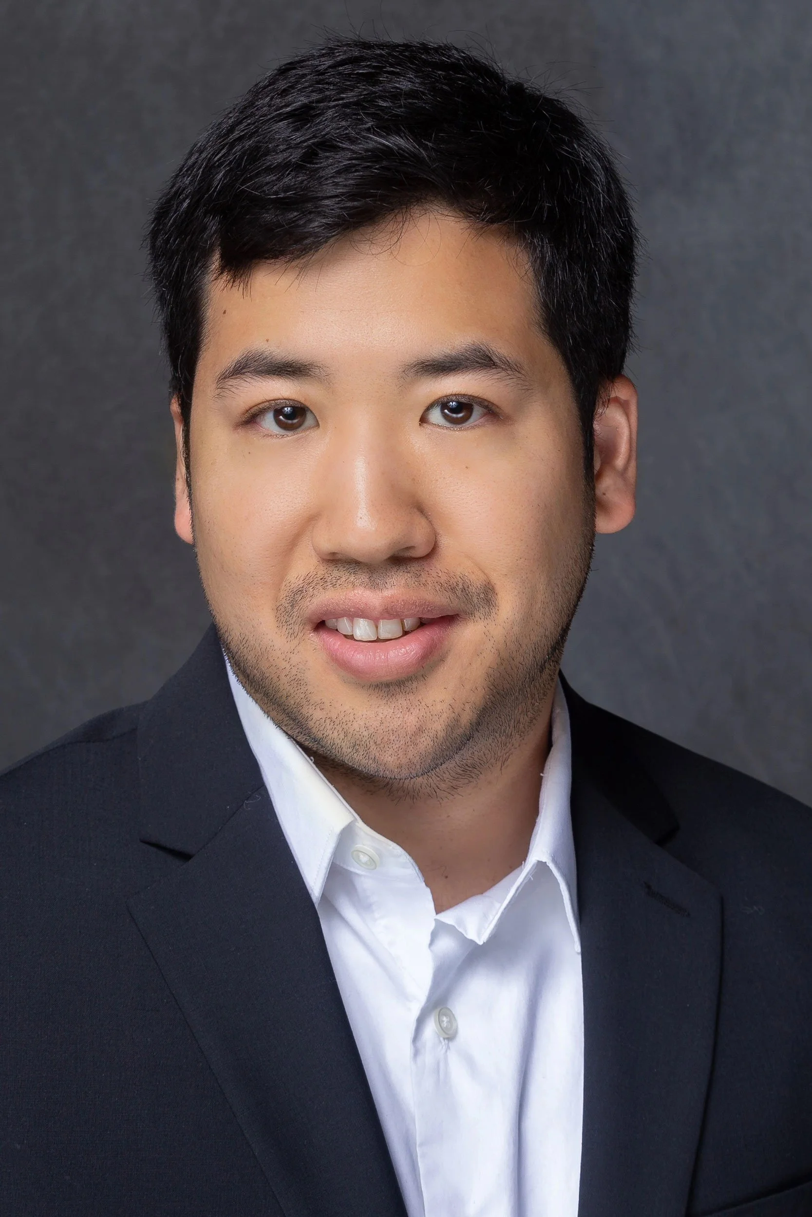 Professional headshot of a young man with black hair, wearing a black suit jacket and white shirt, smiling slightly against a dark gray background.