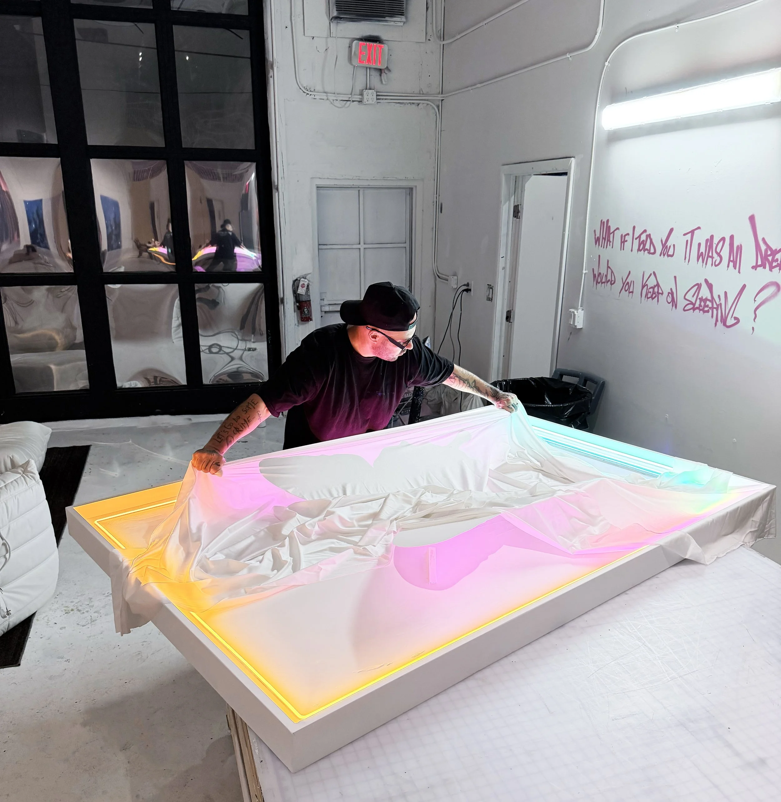 Punkmetender wearing a black cap and shirt is lifting a piece of white fabric from a colorful, illuminated art piece on a table, in a room with industrial decor, a large black window, and pink writing on the wall.