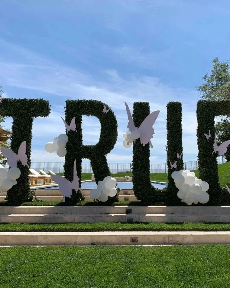 Large decorative letters spelling 'TRUTH' covered in dark greenery, decorated with white balloons and paper butterflies, outdoors on a set of steps with a fountain and grassy area in the background.