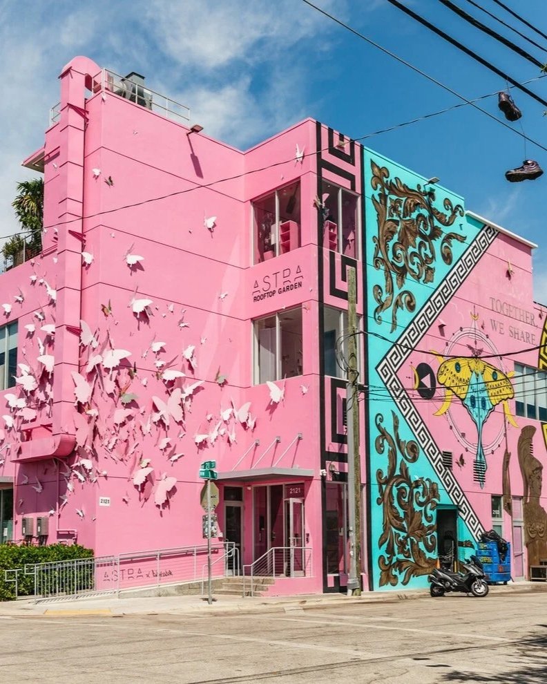 Colorful building with pink and teal walls featuring large butterfly and floral murals, with words 'Astra Rooftop Garden' on pink wall, and pink and white birds sculptures on the pink part.