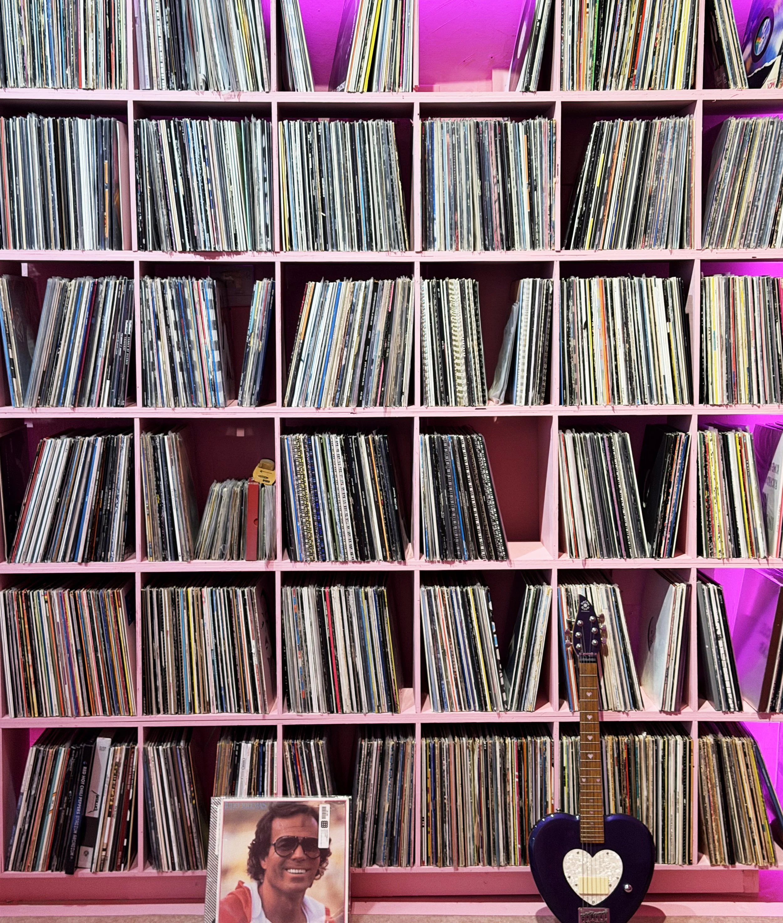 Pink shelving unit filled with vinyl records, a framed photo of a man wearing sunglasses, and a black guitar with a heart-shaped pickguard.
