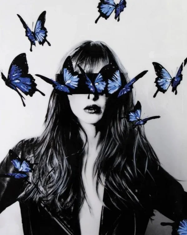 Black and white photo of a woman with long hair, wearing a leather jacket, with blue and black butterflies around her face and some covering her eyes.