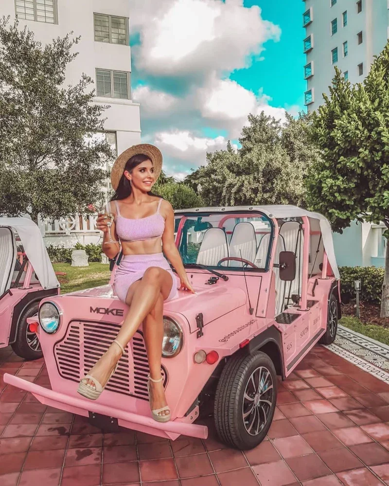 A woman in a pink outfit sitting on the hood of a pink vintage jeep, holding a glass of wine, with trees, buildings, and a partly cloudy sky in the background.
