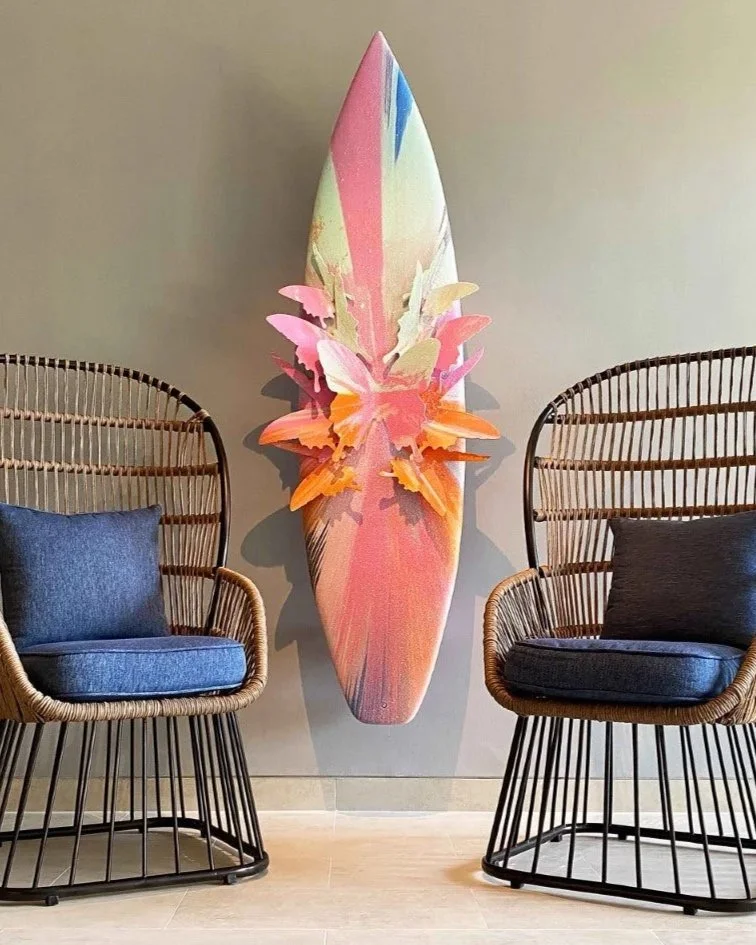 Decorative wall art resembling a surfboard with butterfly-shaped embellishments, placed between two wicker chairs with cushions.