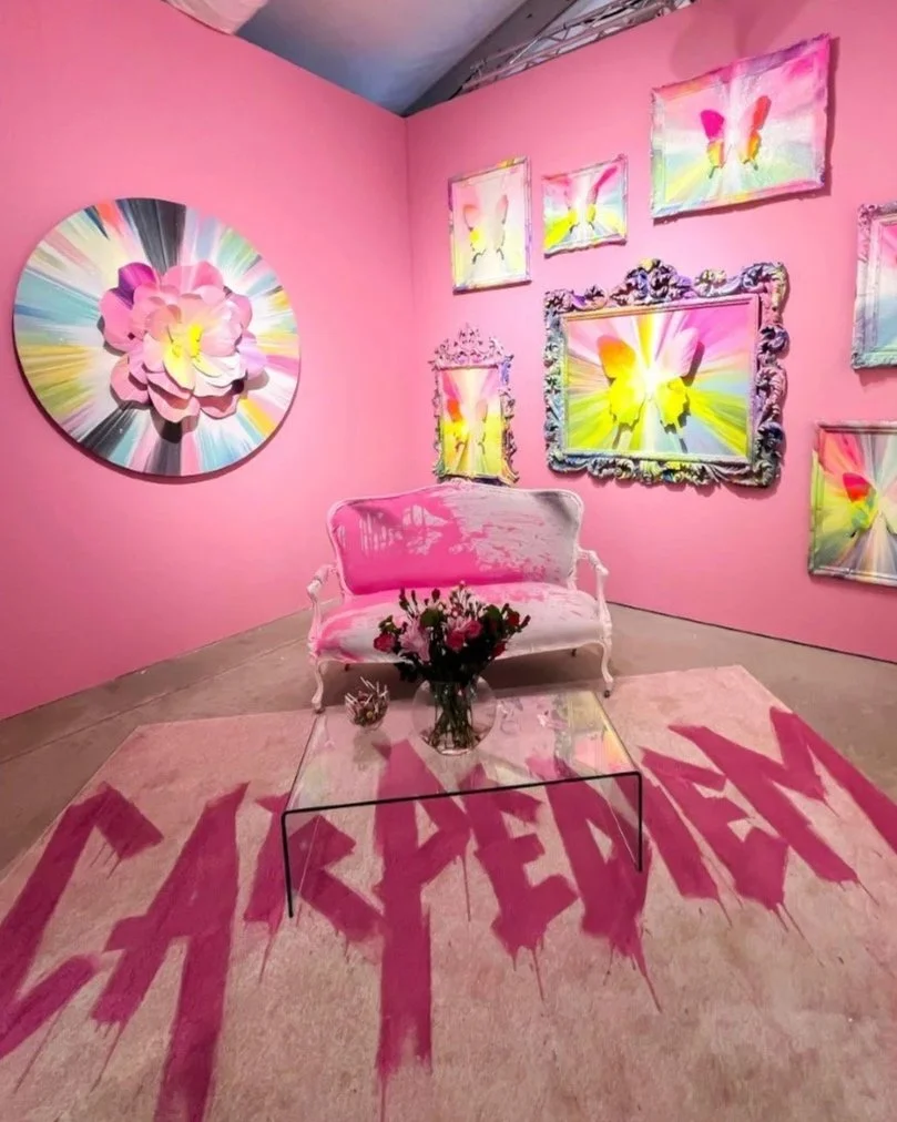 An art gallery with pink walls decorated with colorful floral and butterfly paintings. A white vintage-style sofa with pink brushstroke-like paint on the upholstery is positioned in front of a glass coffee table holding a vase of flowers. A pink rug with the word 'CREATIVE' painted in bold, jagged letters covers the floor.