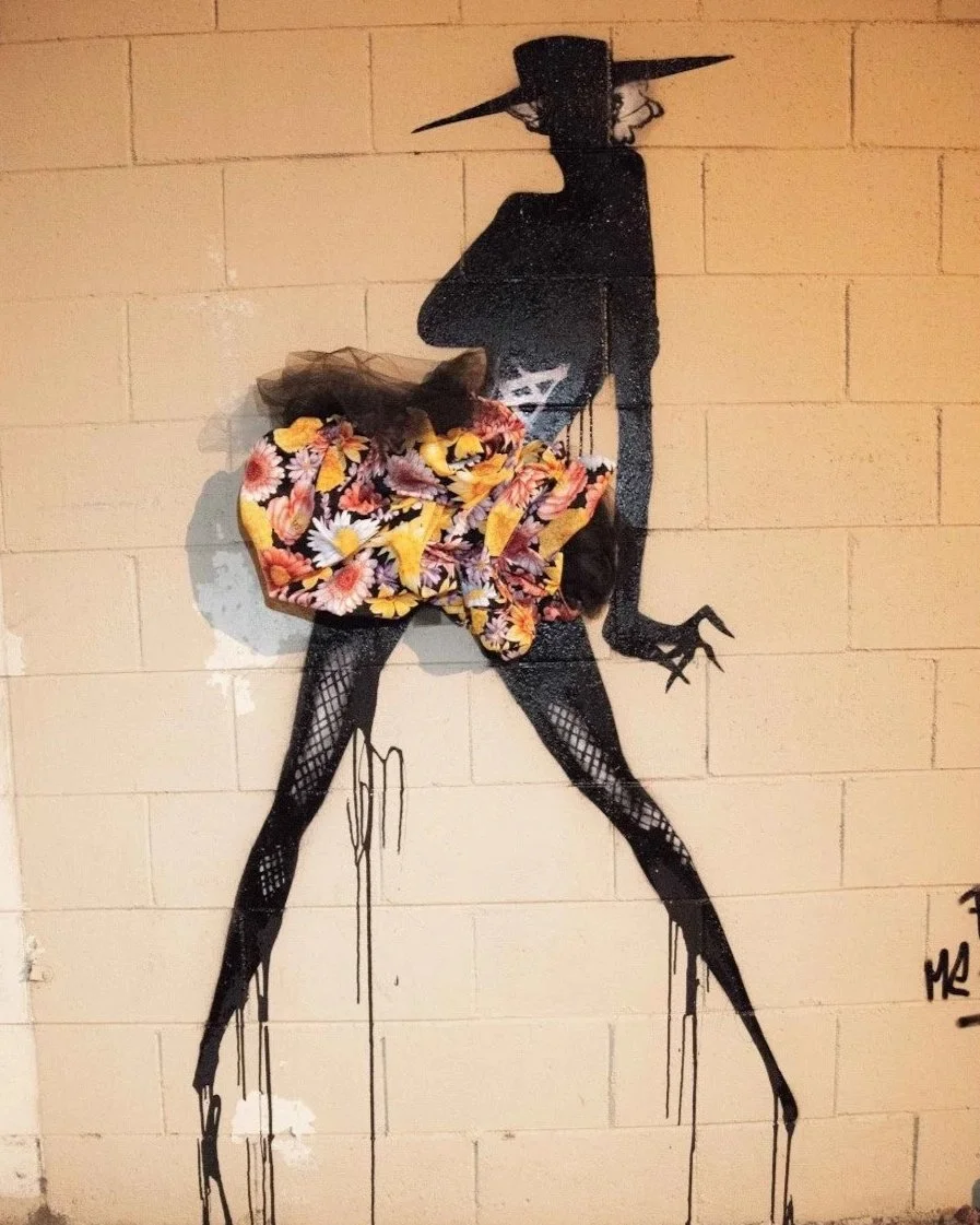 Street art of a silhouette of a woman wearing a hat, decorated with a colorful fabric around the waist, painted on a brick wall.