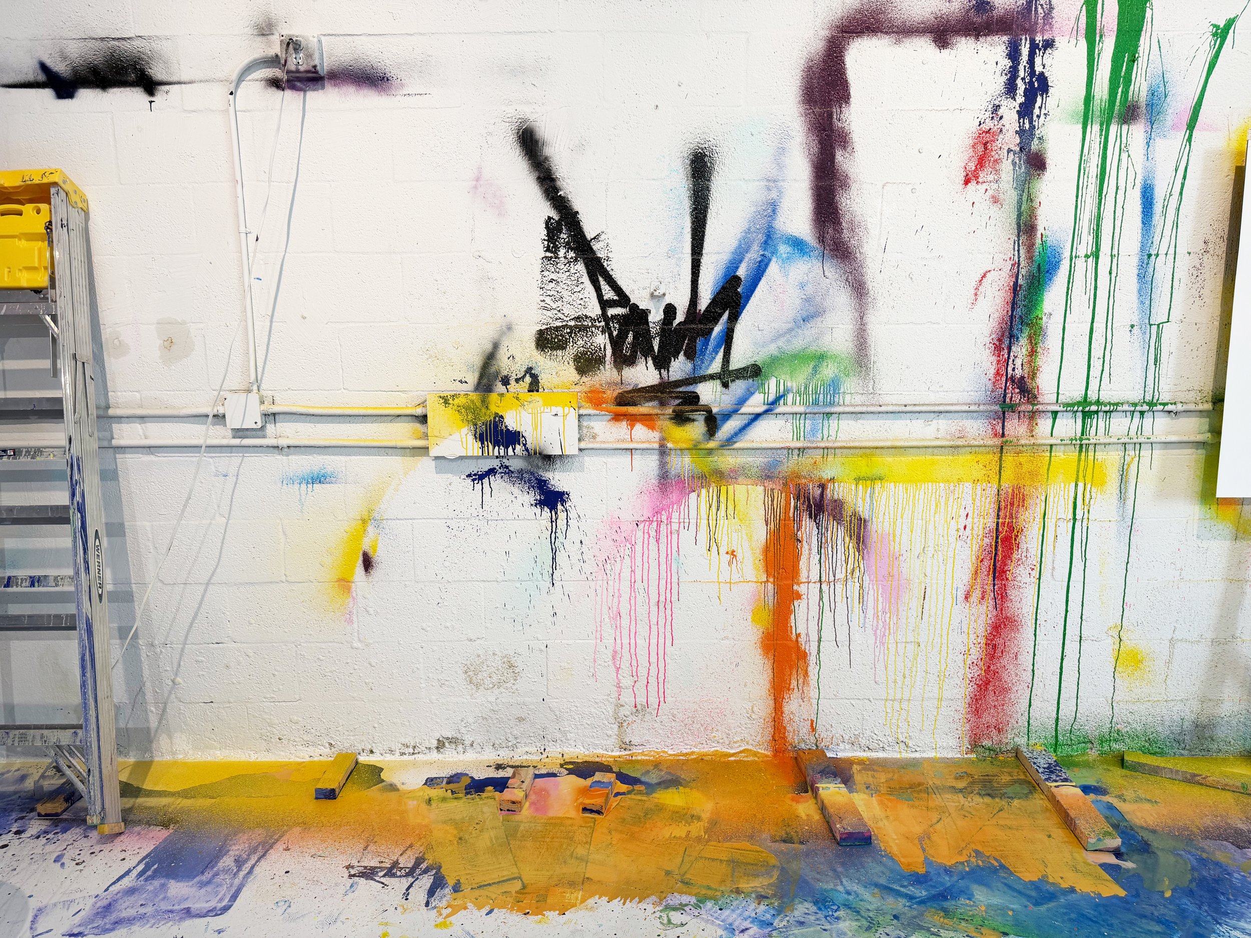 A white wall with colorful spray paint splashes and drips, including black, blue, green, yellow, and red. There is a yellow electrical box and a silver stepladder on the left, and the floor is covered with paint splatters and several wooden blocks.