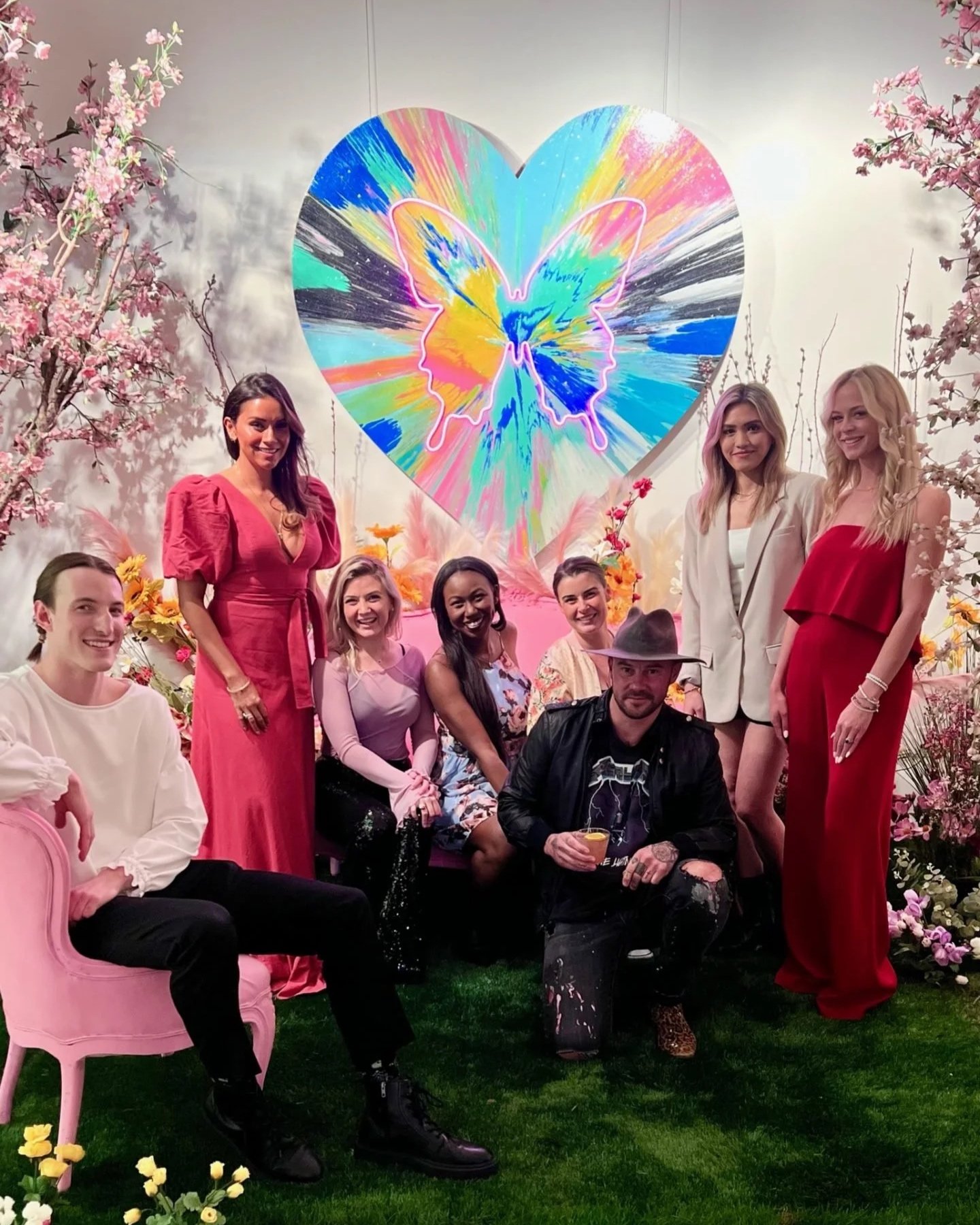 Group of seven people posing in front of colorful art featuring a butterfly outline within a vibrant heart shape, surrounded by flowers and pink blossoms.