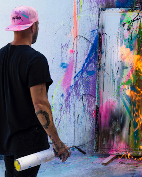 A man wearing a pink cap and black t-shirt spray painting colorful graffiti on a wall and door.