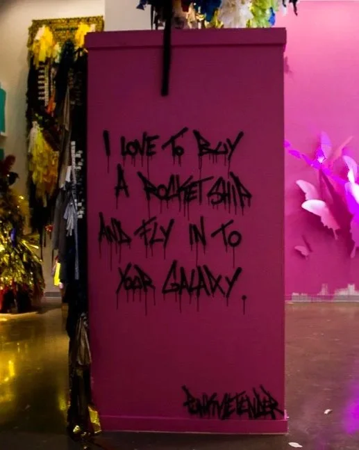 Pink wall with black graffiti text that reads, "I love to buy a rocket ship and fly into your galaxy," signed by someone named "D. Somewhere."