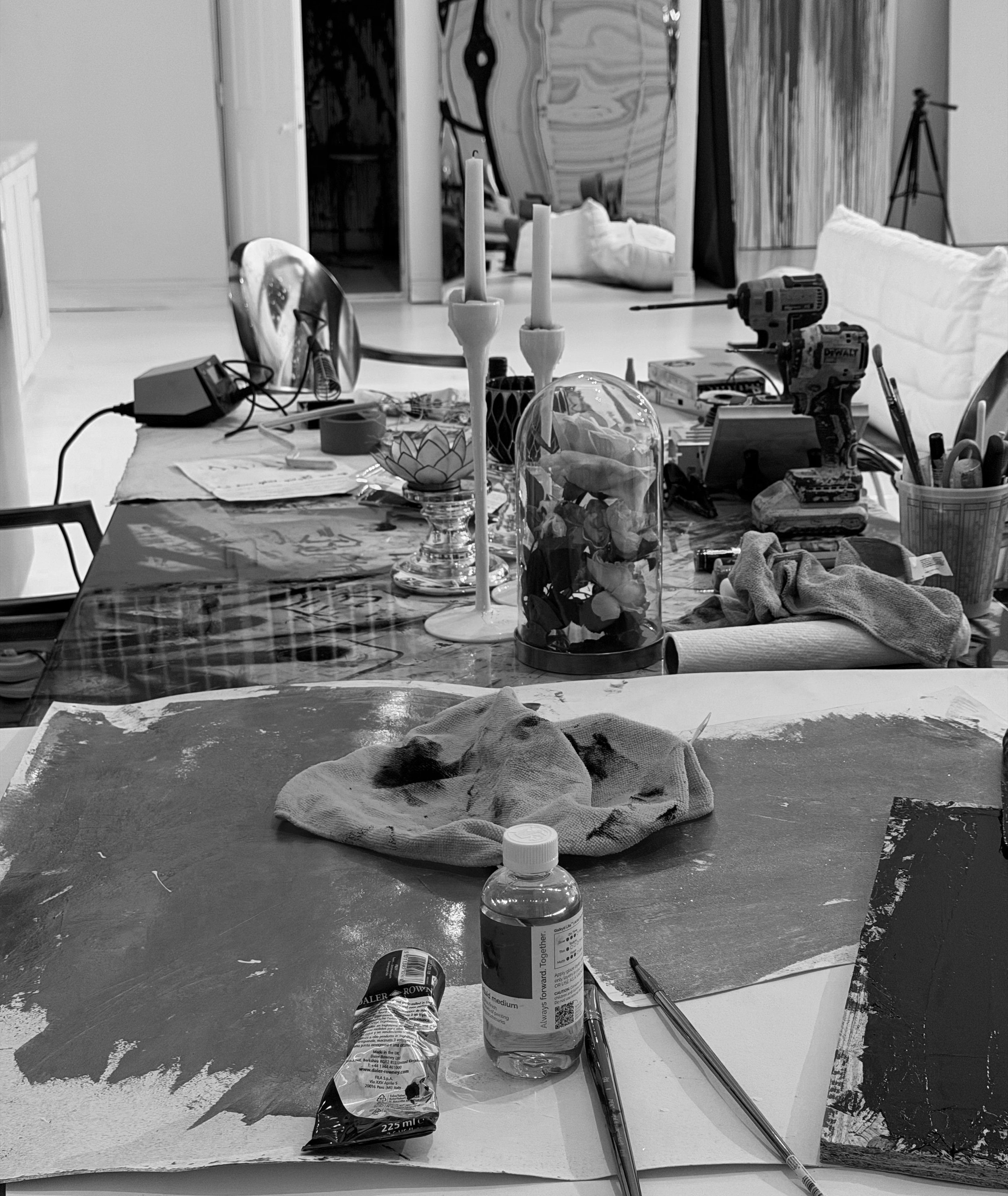 A cluttered dining table with art supplies, tools, candles, a glass-encased flower arrangement, and various other objects in a room with artwork and furniture.