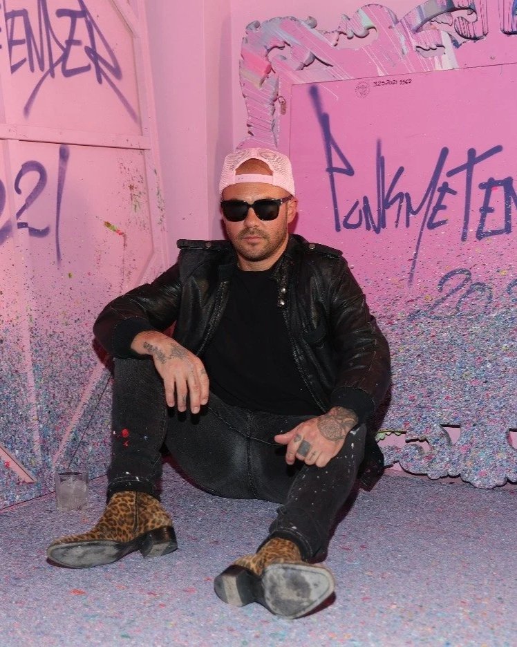 A man sitting on the ground against a pink graffiti-covered wall, wearing a black leather jacket, black pants with paint splatters, leopard print boots, sunglasses, a pink cap backwards, and sporting tattoos.